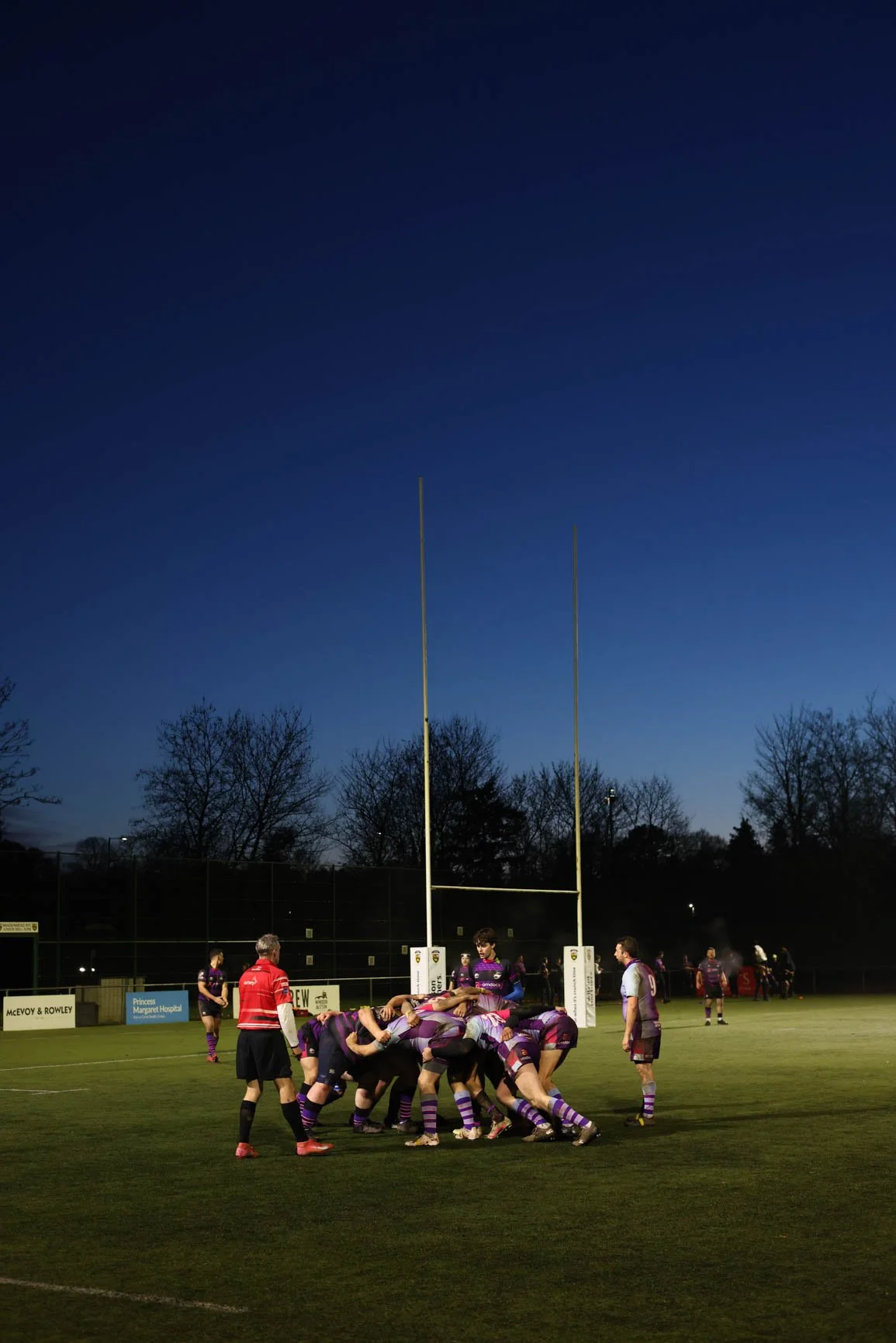 A rugby game is happening on an outdoor field at dusk, with players in purple and white uniforms engaged in a scrum, and a referee nearby.
