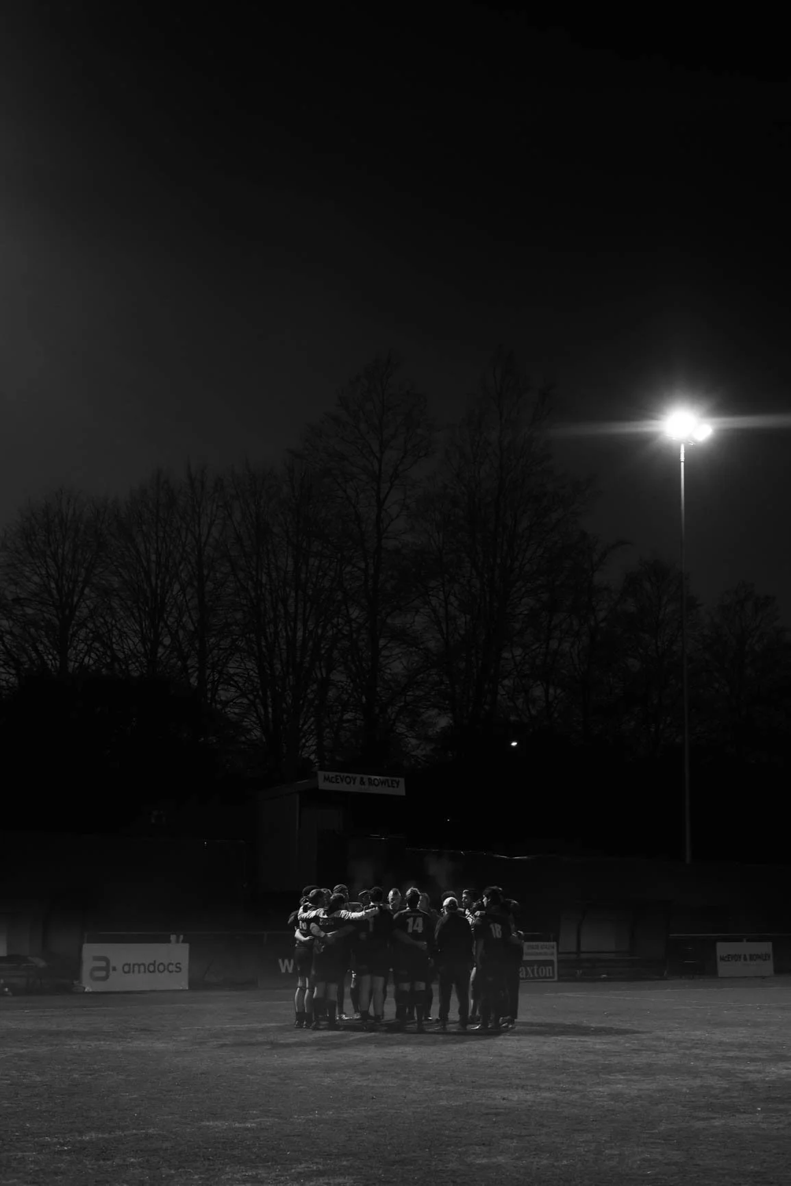 A group of young football players huddled together on a football field at night, illuminated by a large stadium light, with trees in the background.