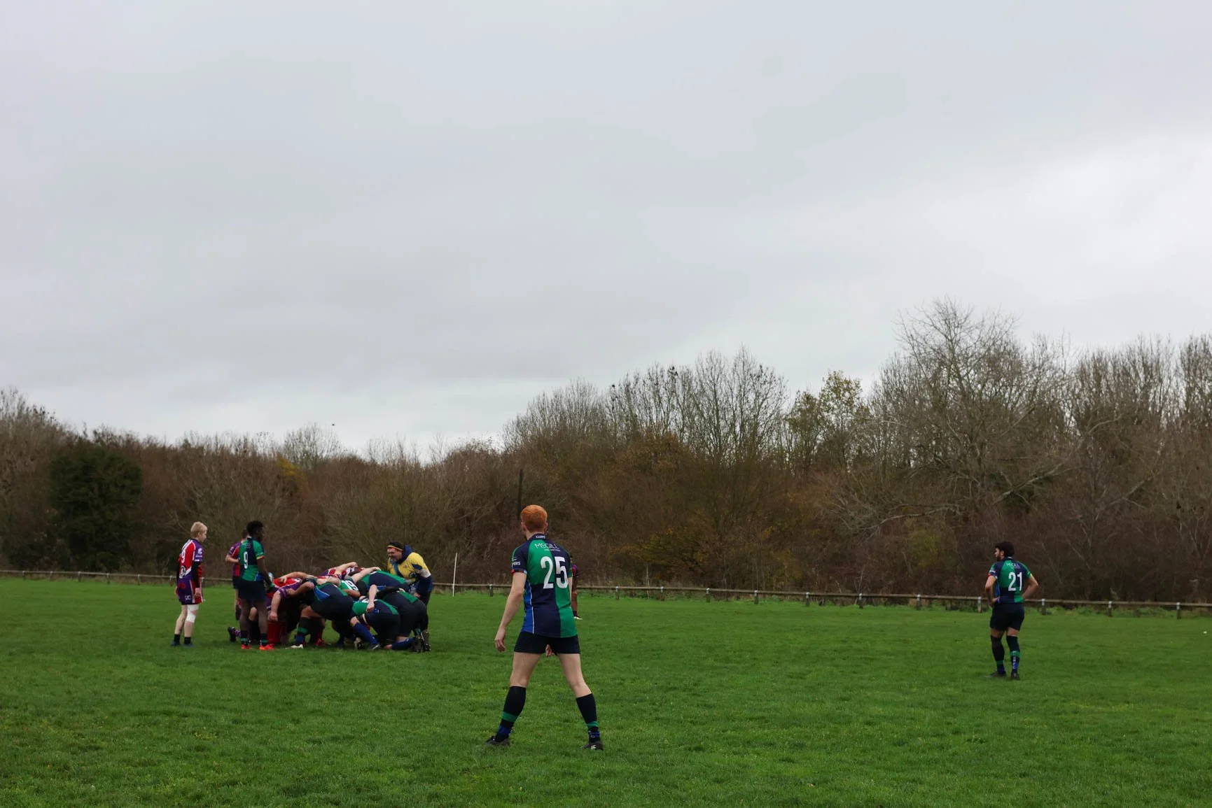 Rugby players engage in a scrum on a grassy field during an overcast day, with some players standing and others crouched down, in a game surrounded by leafless trees.