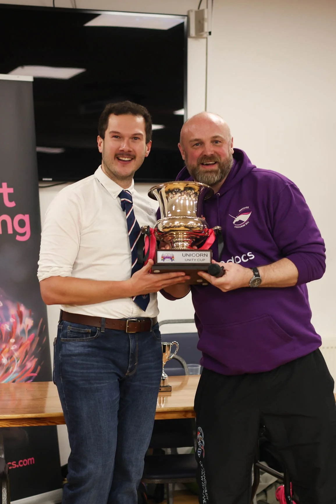 Two men holding a trophy that reads 'Unicorn Unity Cup,' smiling and posing for a photo indoors.