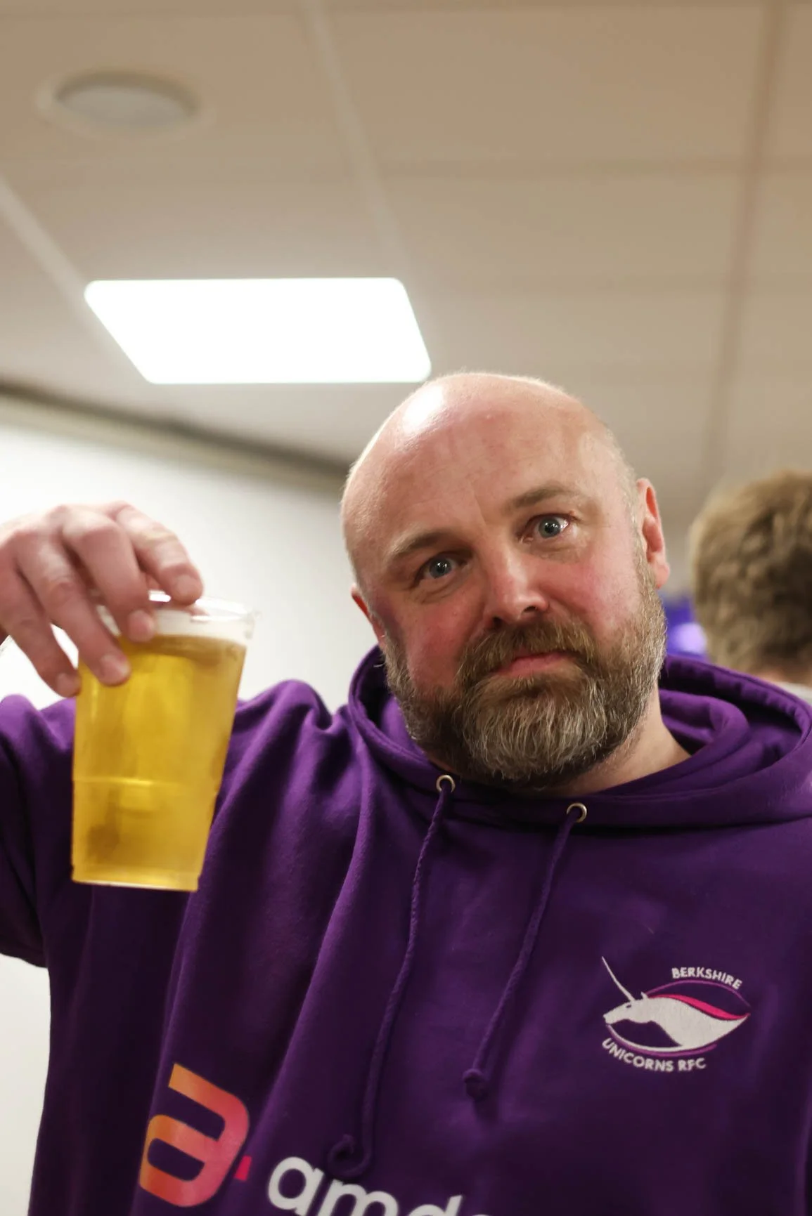 A man with a bald head and beard, wearing a purple hoodie with a logo for Berkshire Unicorns RFC, holding a clear plastic cup filled with a yellow beverage, possibly beer, in a room with a ceiling light fixture.