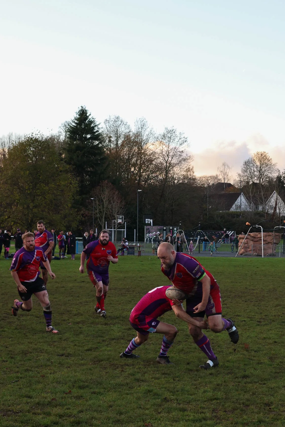 Men playing rugby on a grassy field with a playground and trees in the background, during late afternoon or early evening.