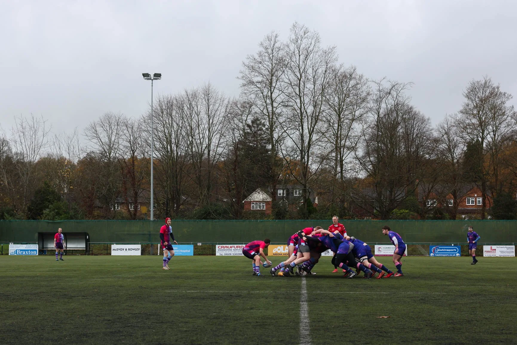 A rugby match in progress on a grass field with players engaged in a scrum, surrounded by a green fence and advertising boards. Several houses and leafless trees are visible in the background on an overcast day.