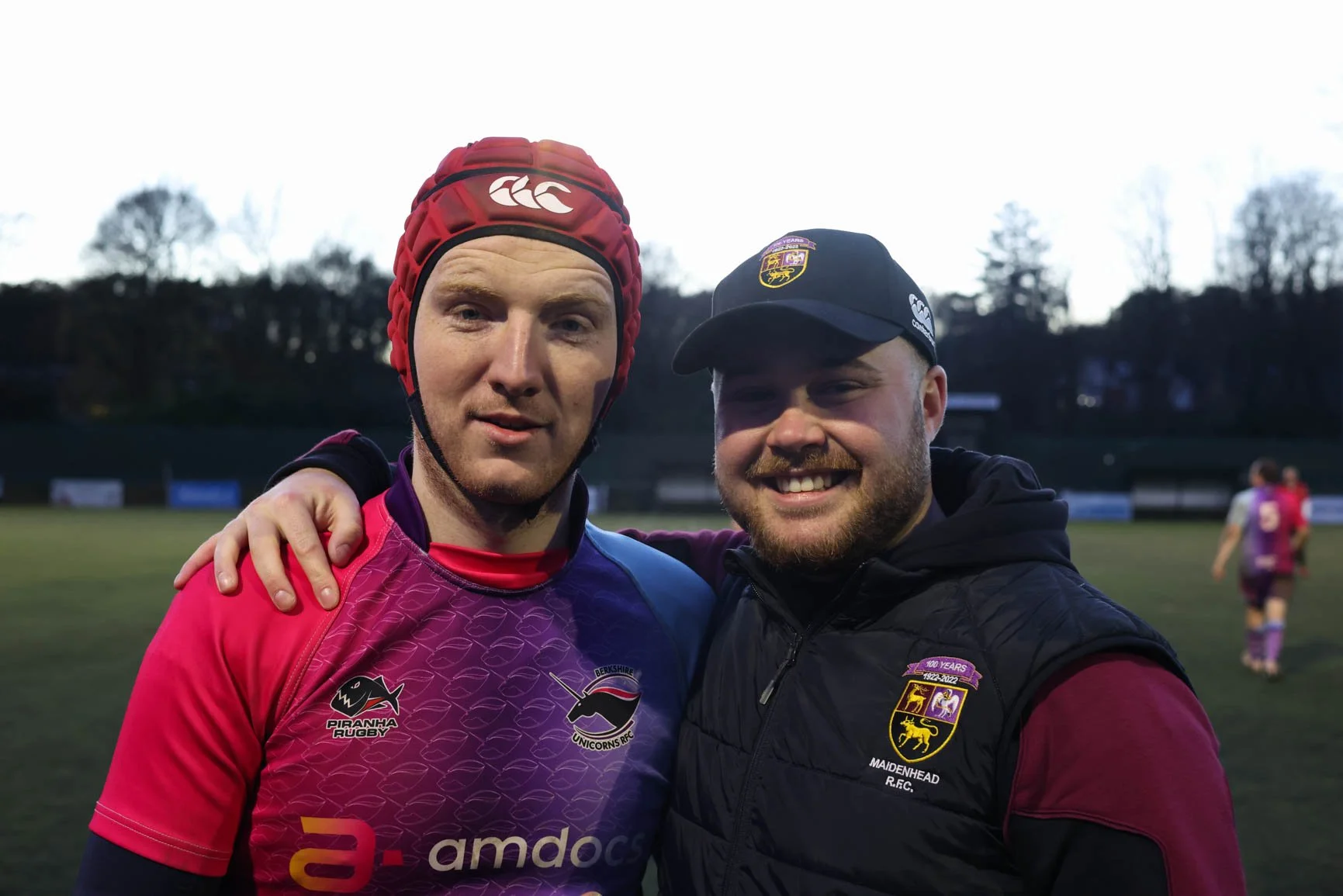 Two smiling men posing together on a sports field in the evening, wearing rugby attire and hats, with others playing in the background.
