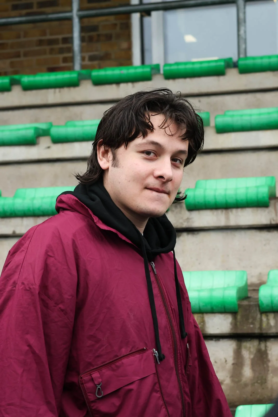 A young man with dark wavy hair wearing a maroon jacket with a black hoodie underneath, standing outdoors in front of concrete steps with green railings.