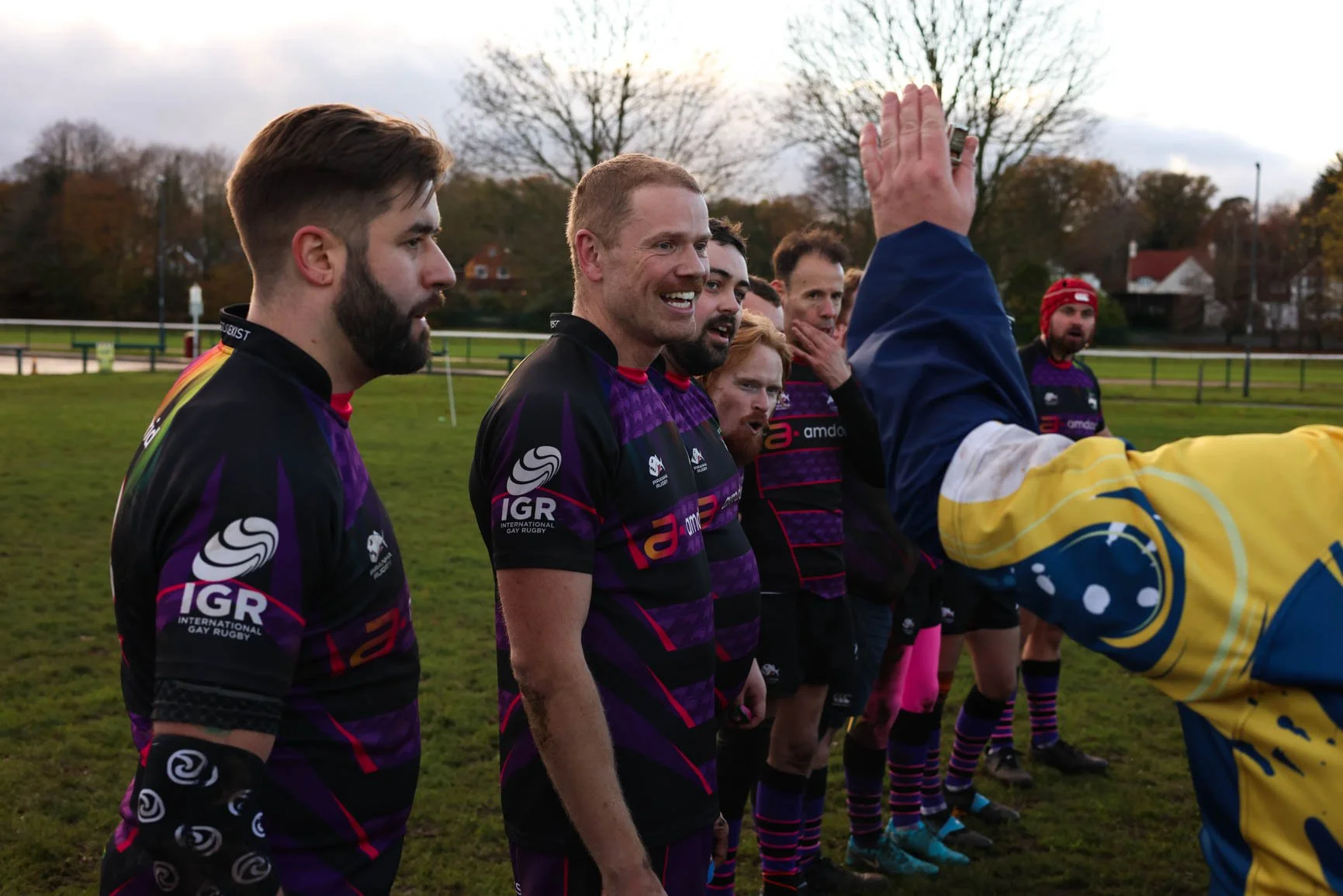A group of rugby players in purple and black uniforms standing on a grass field, receiving instructions from a coach in a yellow and blue jacket. The players are outdoors with trees and houses in the background under an overcast sky.