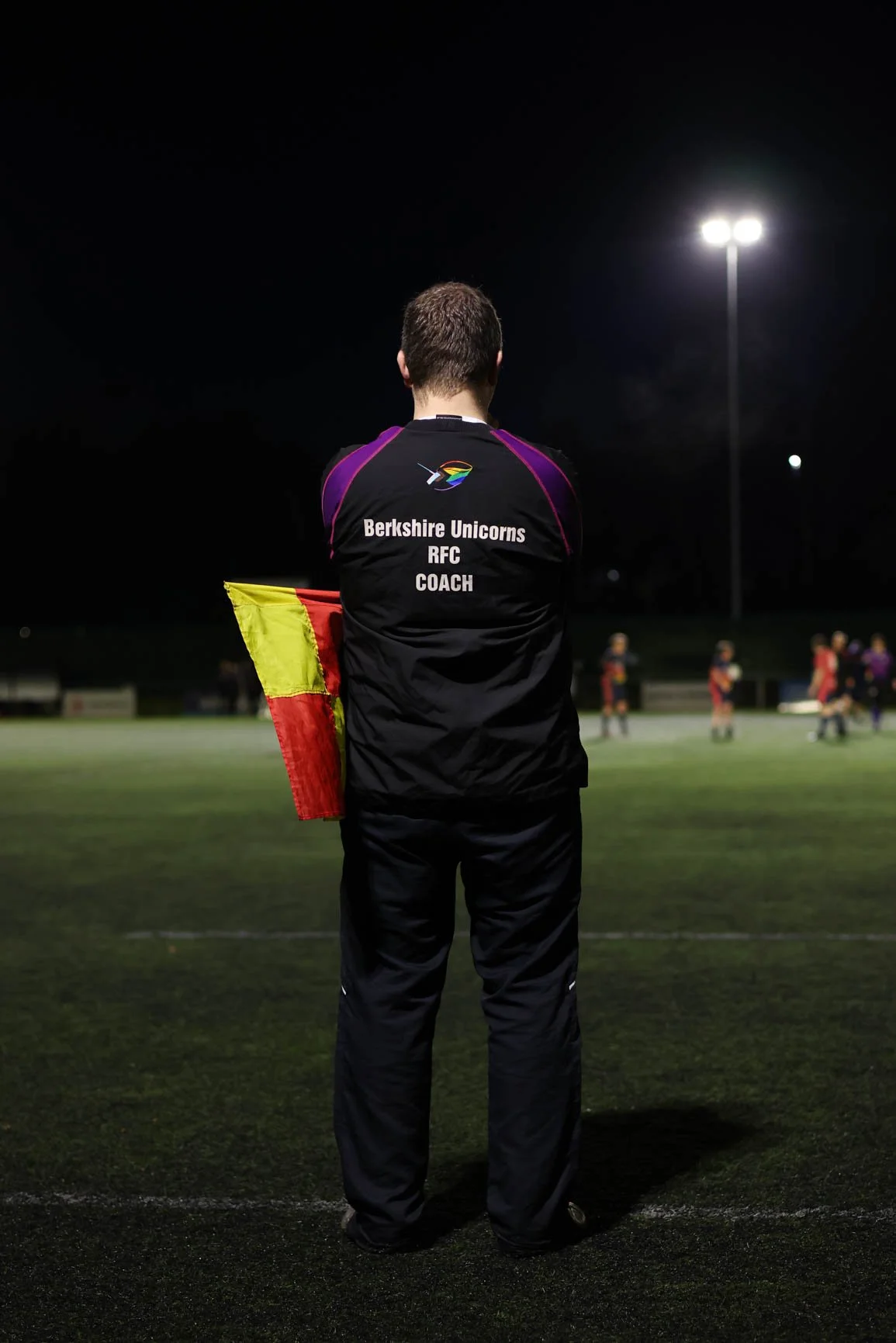 A soccer coach standing on a field at night, holding a yellow and red flag, with players visible in the background under stadium lights.