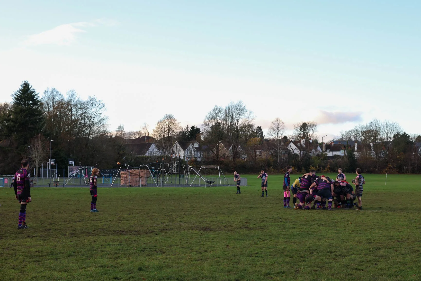 Rugby players on a field during a game, with some players scrummaging and others standing around, and a playground with slides and swings in the background near houses and trees.