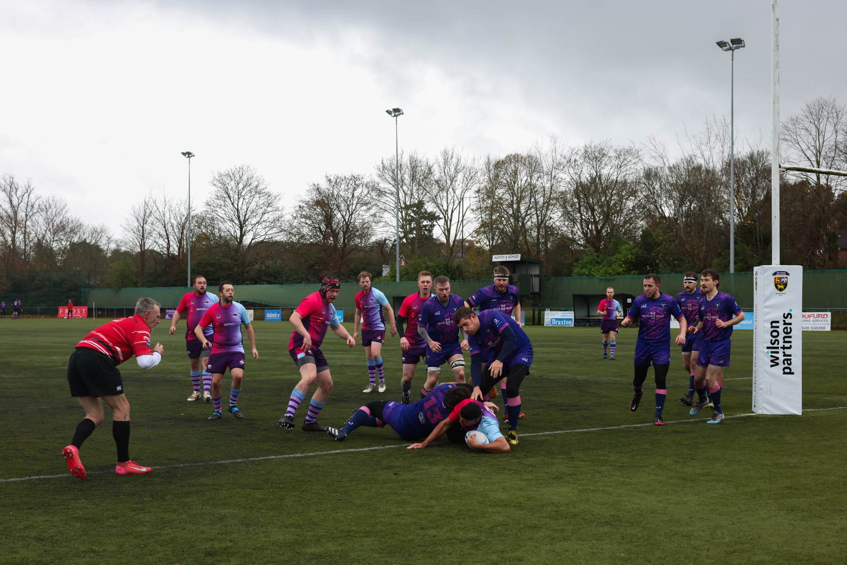 Rugby players in purple and red jerseys engaged in a match on a grass field, with several players tackling a player holding the rugby ball.