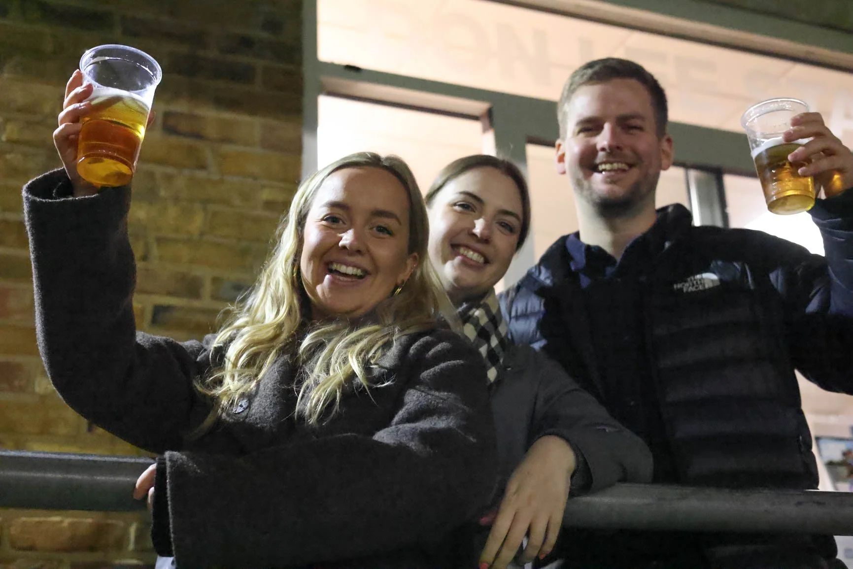 Three people smiling and holding up cups of beer outdoors at night in front of a brick wall and a lit window.