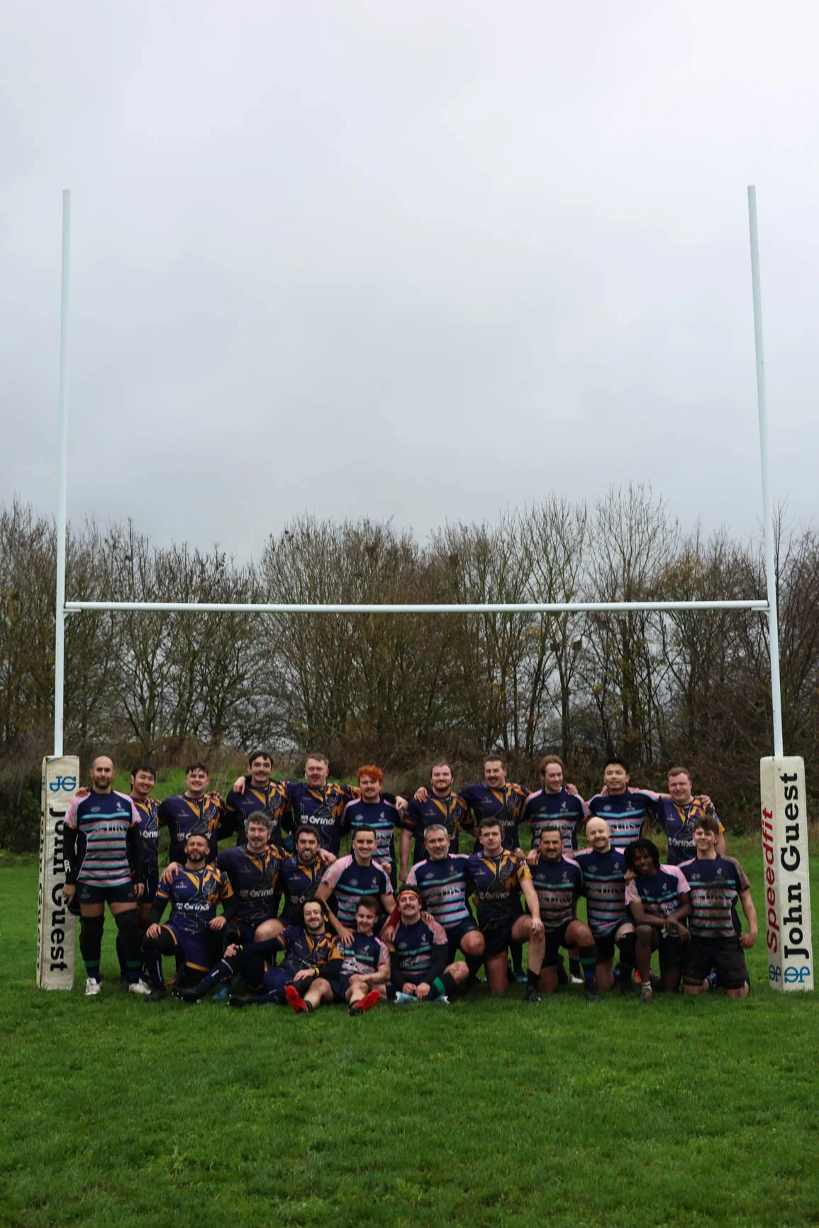 A group of rugby players poses on a field under goalposts, wearing matching jerseys, with trees and an overcast sky in the background.