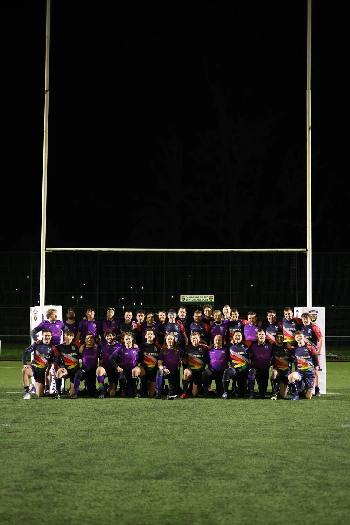 Group of rugby players in purple and black uniforms posing for a team photo on a rugby field at night, with goalposts and a sign that says 'Maidenhead Rugby Club Junior Skill Zone' in the background.