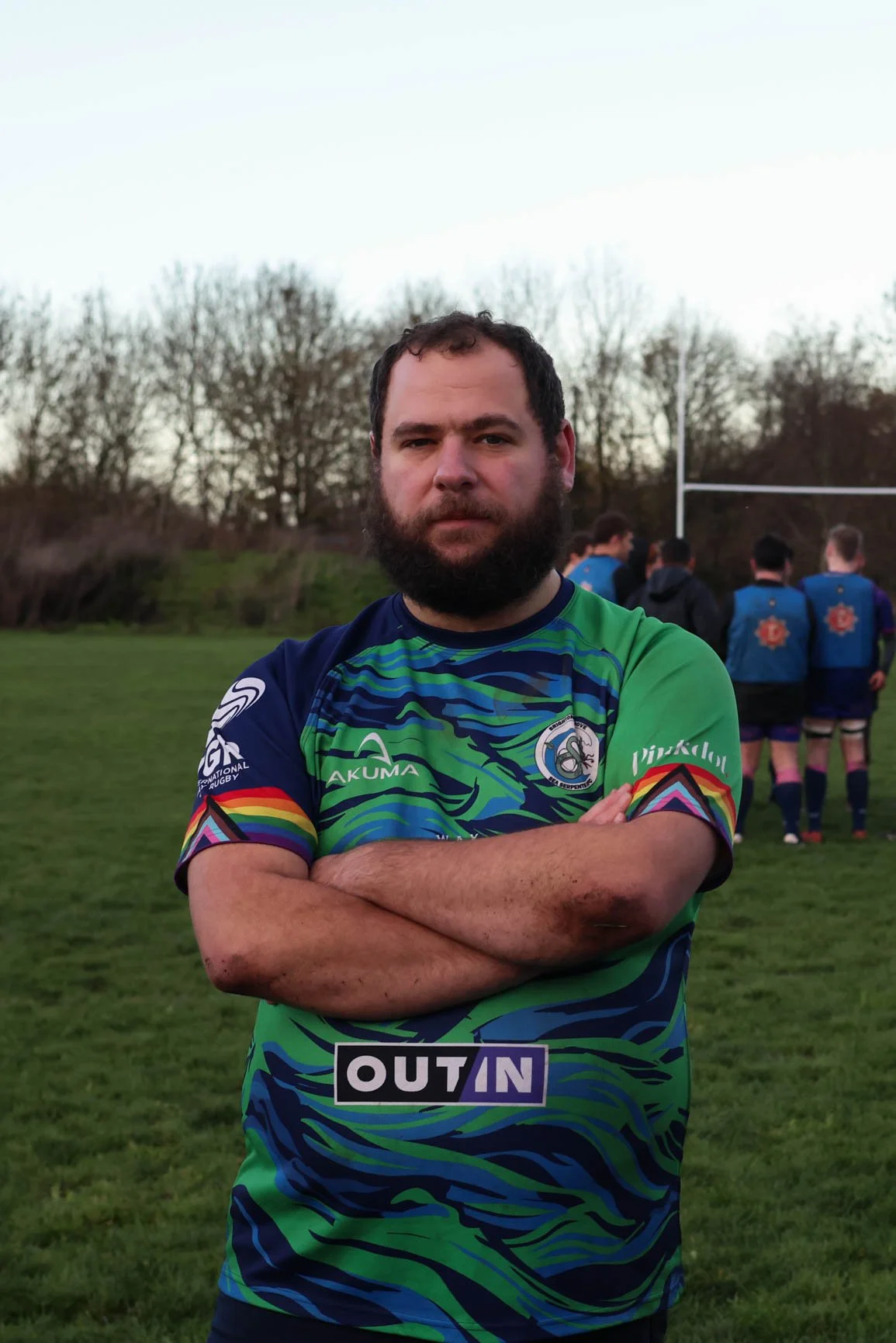 Man with a beard in a colorful rugby jersey standing on a field with crossed arms, with rugby goal posts and other players in the background during sunset.