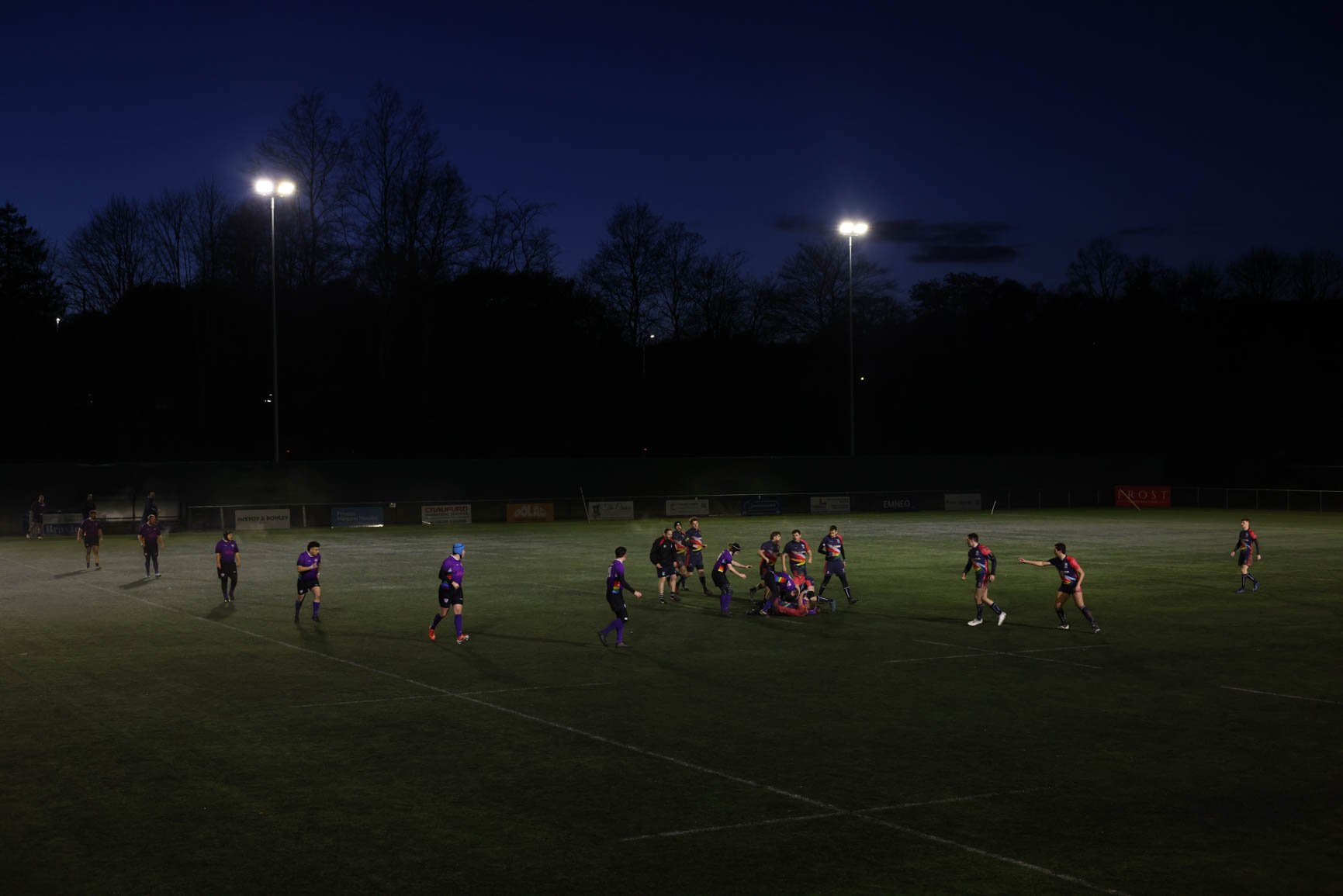 A rugby match is happening under stadium lights at night. Players are on the field, some standing and others gathered around, with one player lying on the ground. The background shows trees and dark sky.