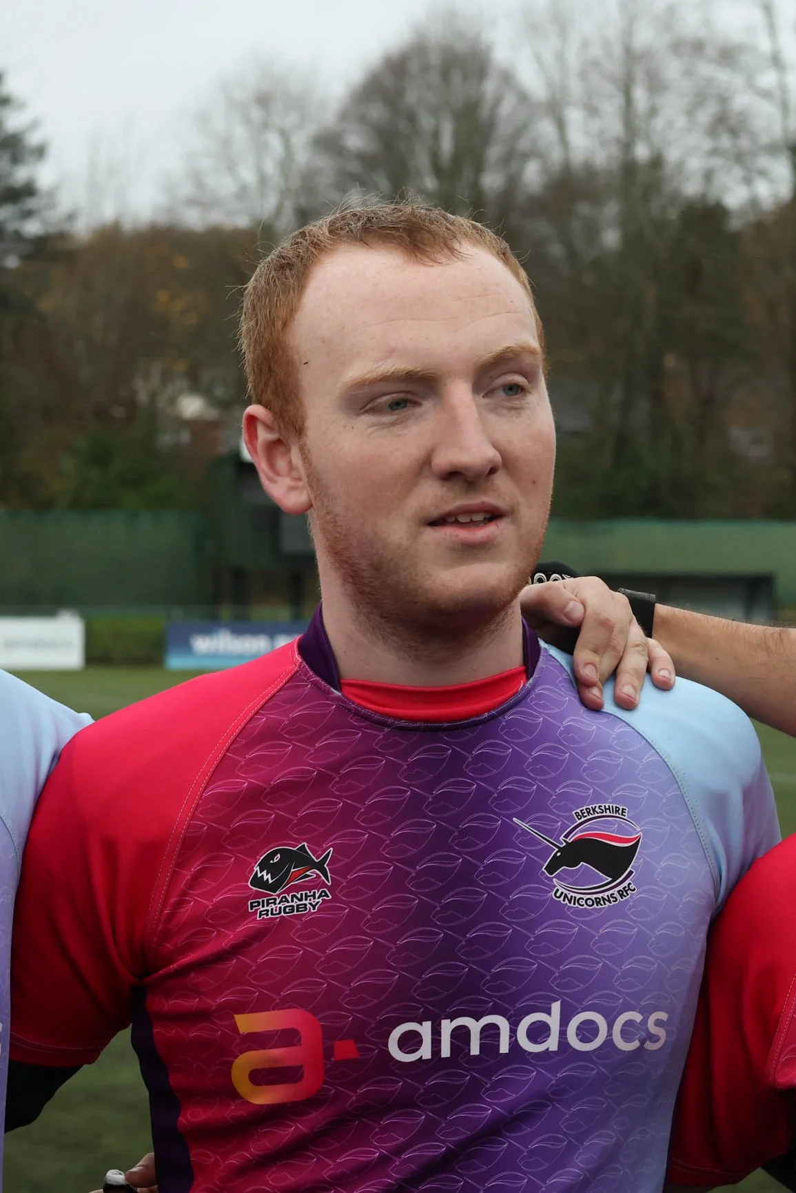 A young man in a rugby uniform standing outdoors on a field with trees in the background, posing with an arm around his teammates.
