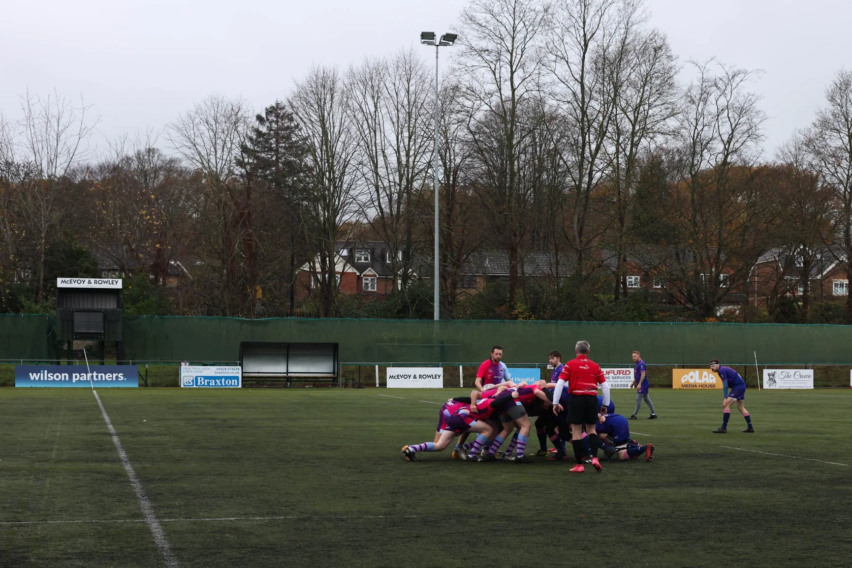 A rugby match in progress on a grass field, with players engaged in a scrum, some in purple and pink uniforms, and referees and spectators visible in the background. Trees and houses are seen beyond the field.