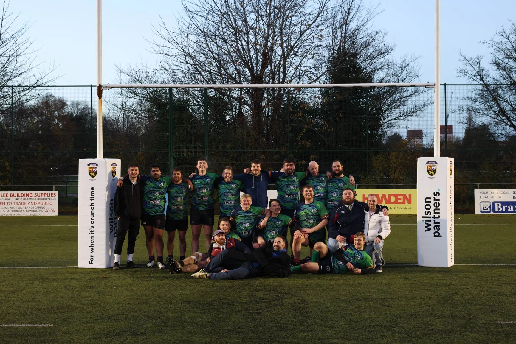 A group of rugby players and staff posing for a team photo on a rugby field, with goalposts and advertising boards in the background, during evening or late afternoon.