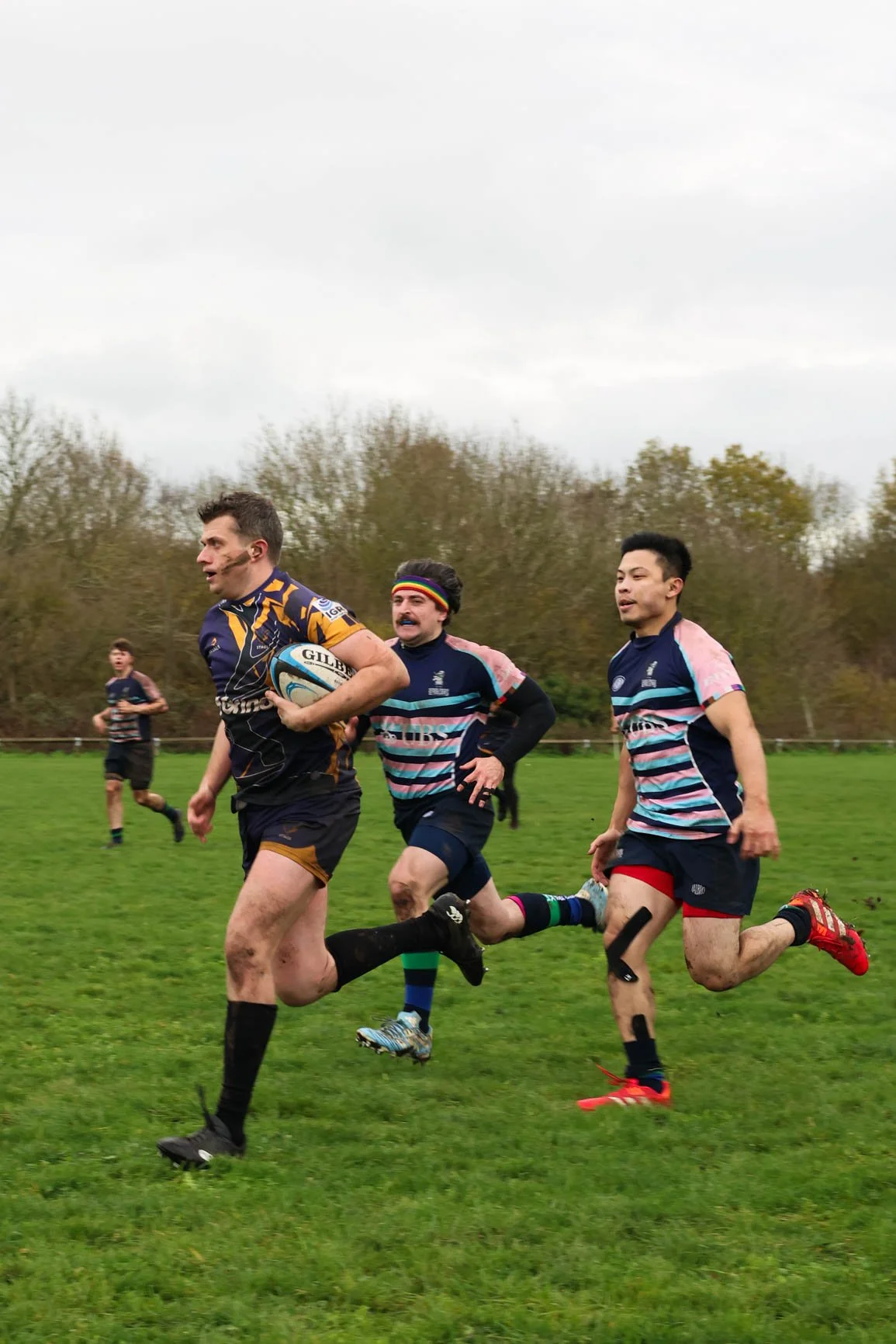 Rugby players running on a grassy field during a match, with one holding a rugby ball, others in motion, and trees in the background.