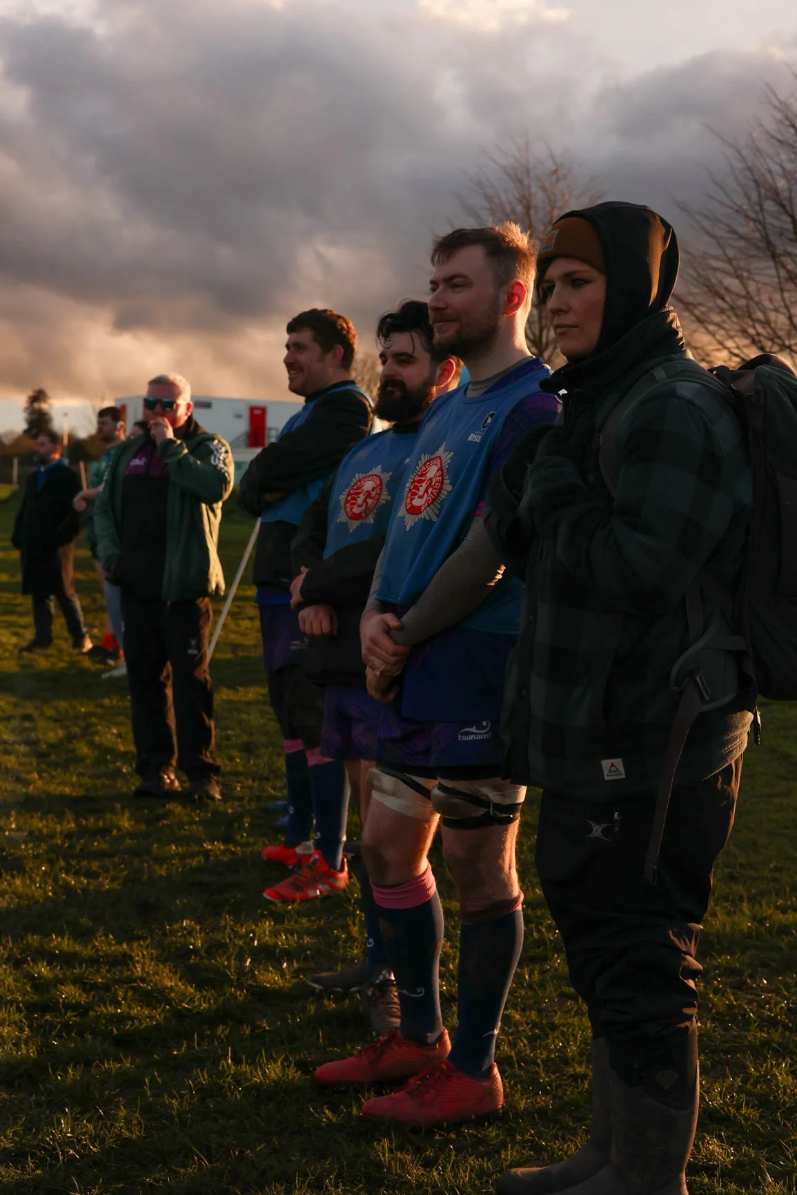 Group of people standing outdoors on grass during sunset, some wearing sports attire and others in jackets, with clouds in the sky.