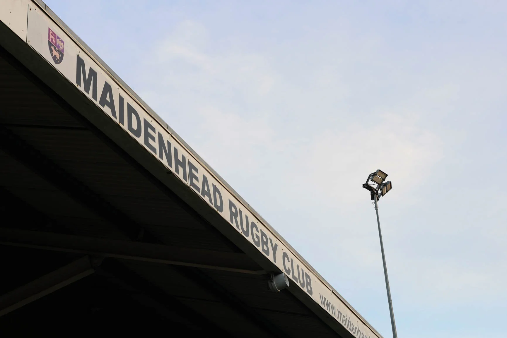 Part of a stadium or sports club sign that reads 'Maidenhead Rugby Club' with the club's crest and a tall floodlight against a cloudy sky.