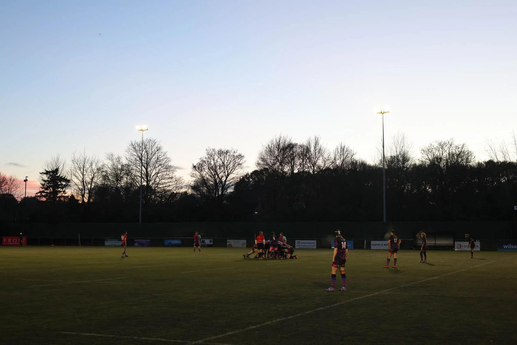 A rugby match taking place at dusk on an outdoor field, with players gathered in a scrum. The background shows a sky transitioning from day to night, with a mix of light and dark clouds, and trees with bare branches. Stadium lights are on, illuminati
