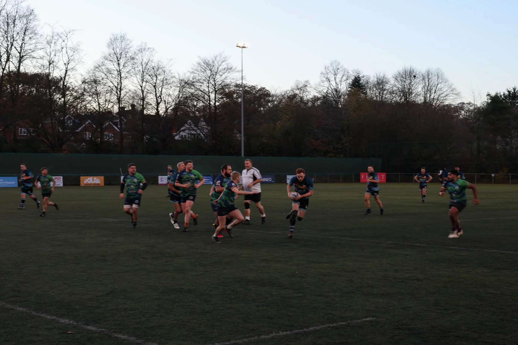 A rugby game being played on a grassy field, with players running and holding a rugby ball, under a clear evening sky, surrounded by trees and houses in the background.