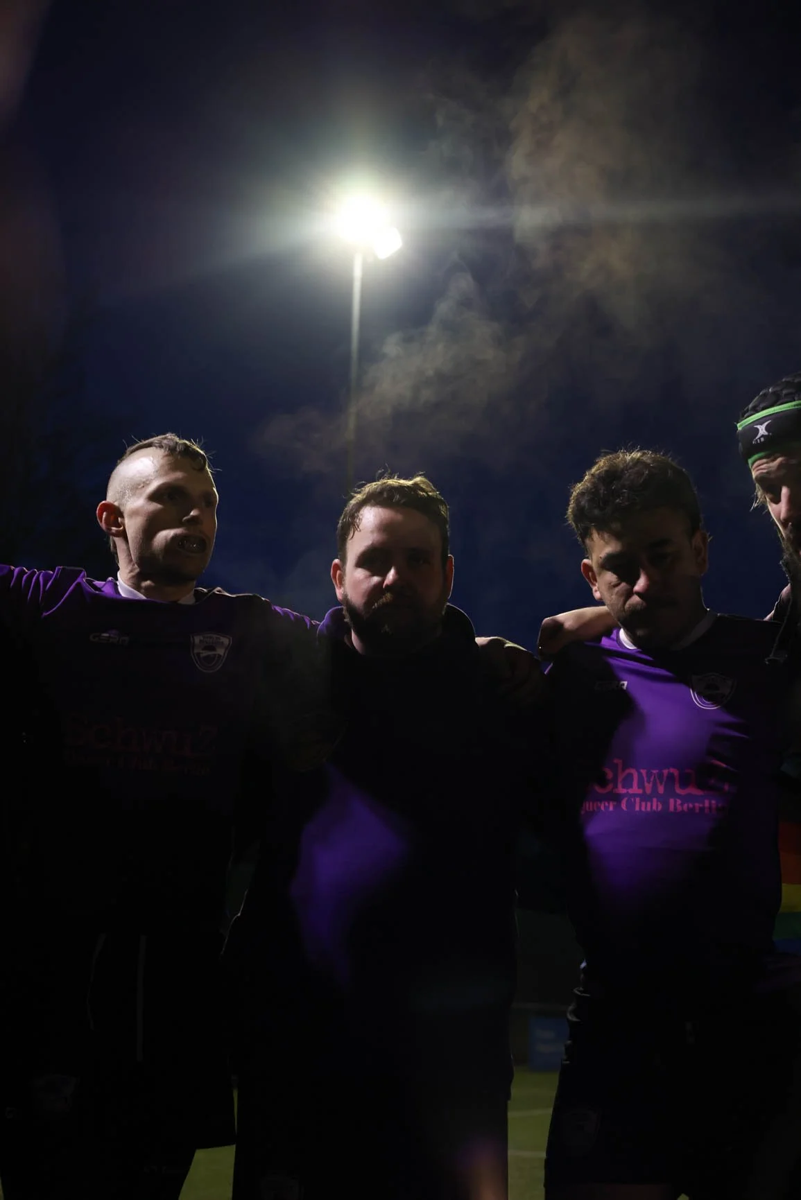 Nighttime photo of five soccer players standing together with arms around each other, illuminated by bright stadium lights against a cloudy sky.