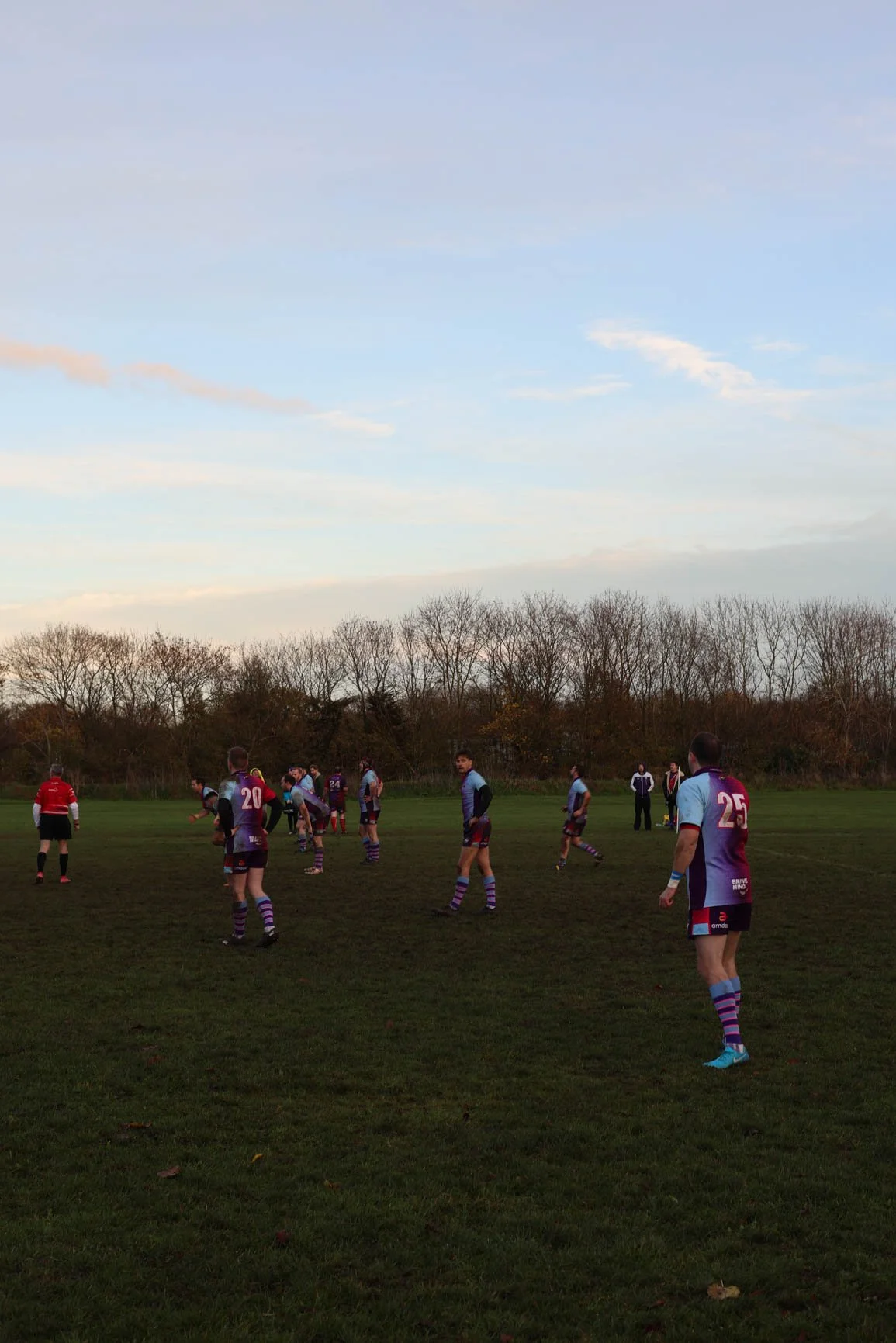 A group of rugby players on a field during late afternoon or early evening. The players are wearing purple and teal uniforms, with some standing and others preparing for play. The background features trees with autumn foliage under a partly cloudy sk
