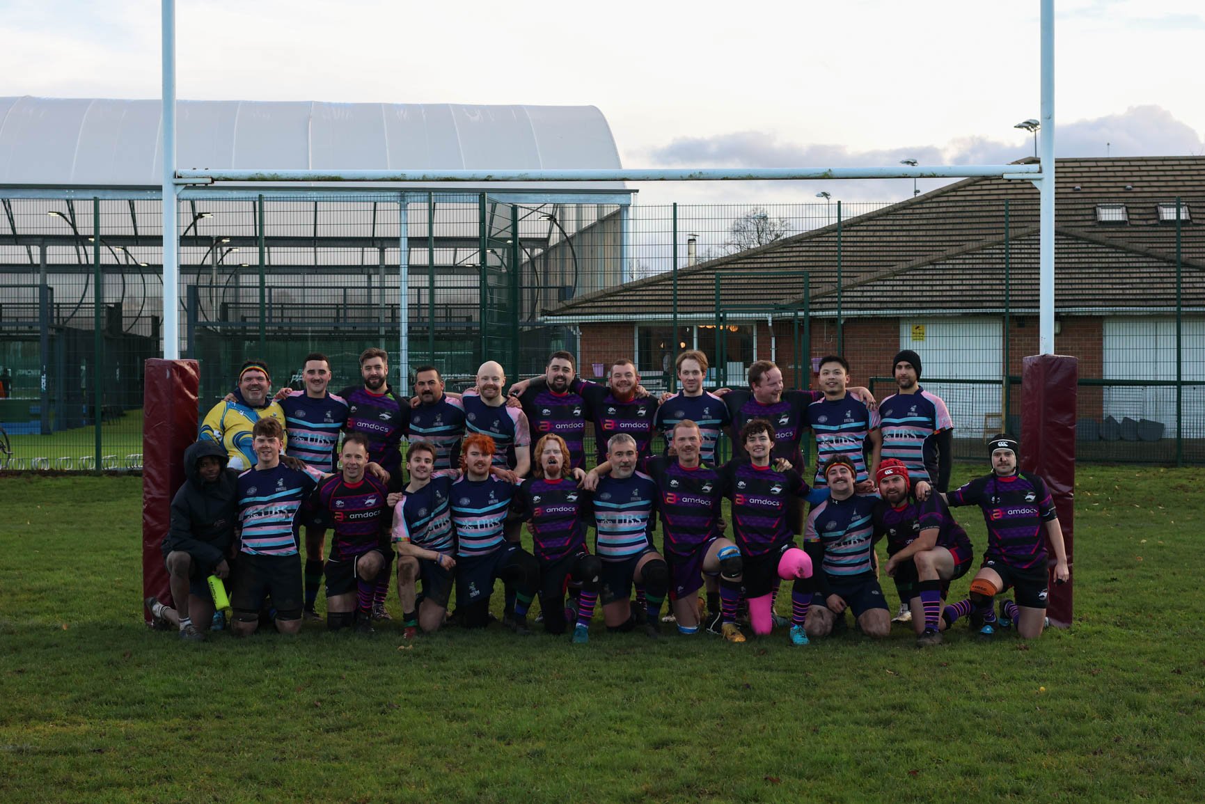A group of rugby players and coaches is standing on a rugby field in front of a rugby goalpost, posing for a team photo. They are wearing matching dark jerseys with colorful stripes and logos, and some are wearing headgear and knee pads. The backgrou