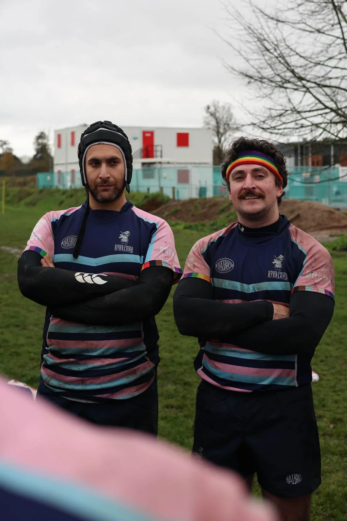 Two male rugby players standing outside on a cloudy day, wearing colorful team jerseys with the word 'STEELERS' and a crest, both with arms crossed, one wearing a rugby helmet and the other a rainbow headband, in front of a grassy field and some tree