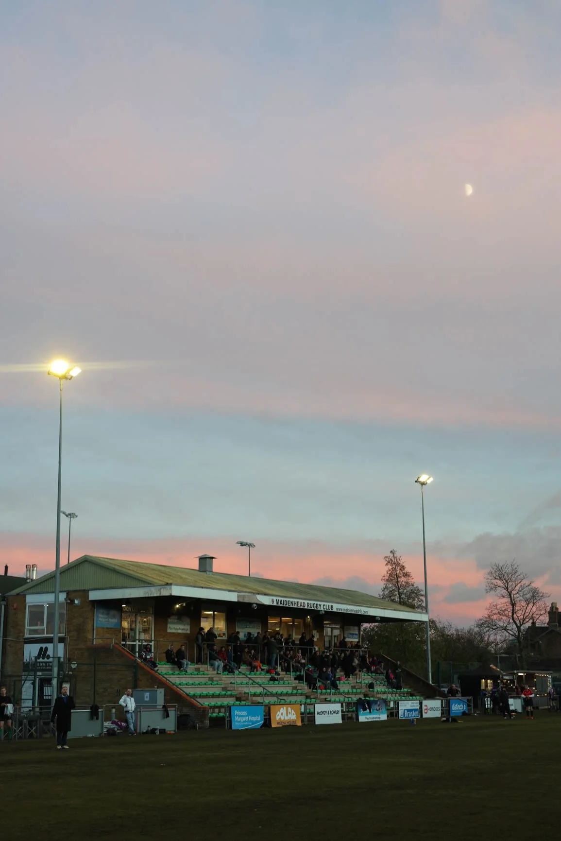 Rugby club building with spectators in stands, outdoor field, evening sky with clouds, moon, and tall stadium lights