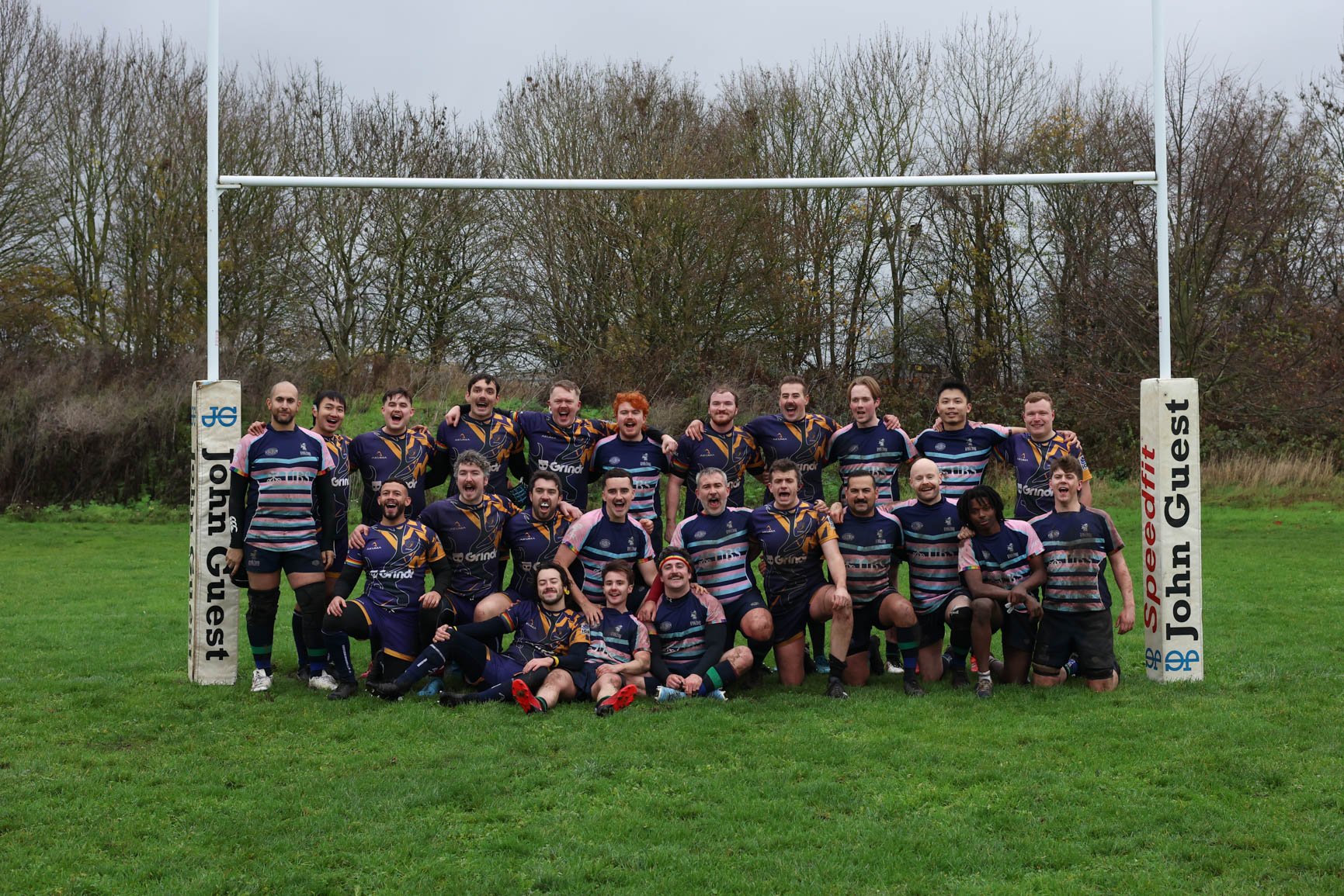 A group of rugby players posing on a field under an overcast sky, with a goalpost behind them and trees in the background.