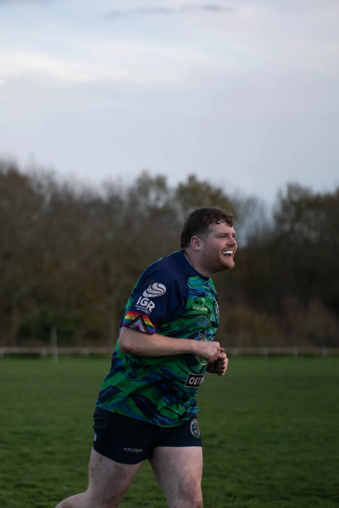 A young man running on a grassy field during twilight, wearing a colorful sports jersey and shorts, smiling and showing enthusiasm.