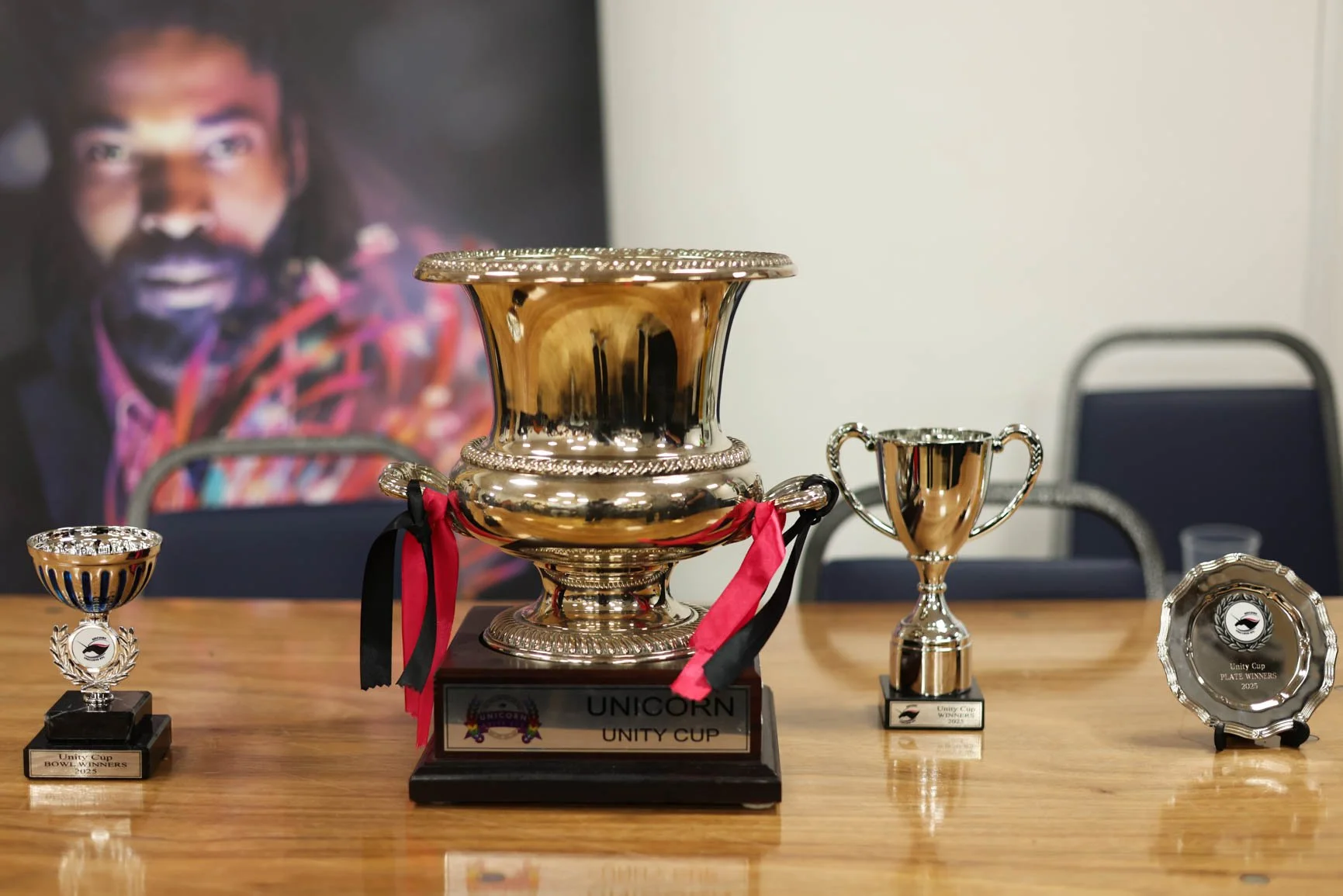 Several trophies and awards on a wooden table, with a large metallic cup in the center, a smaller cup to the right, and two smaller awards on either side, behind a sign that reads 'Unicorn Unity Cup'.