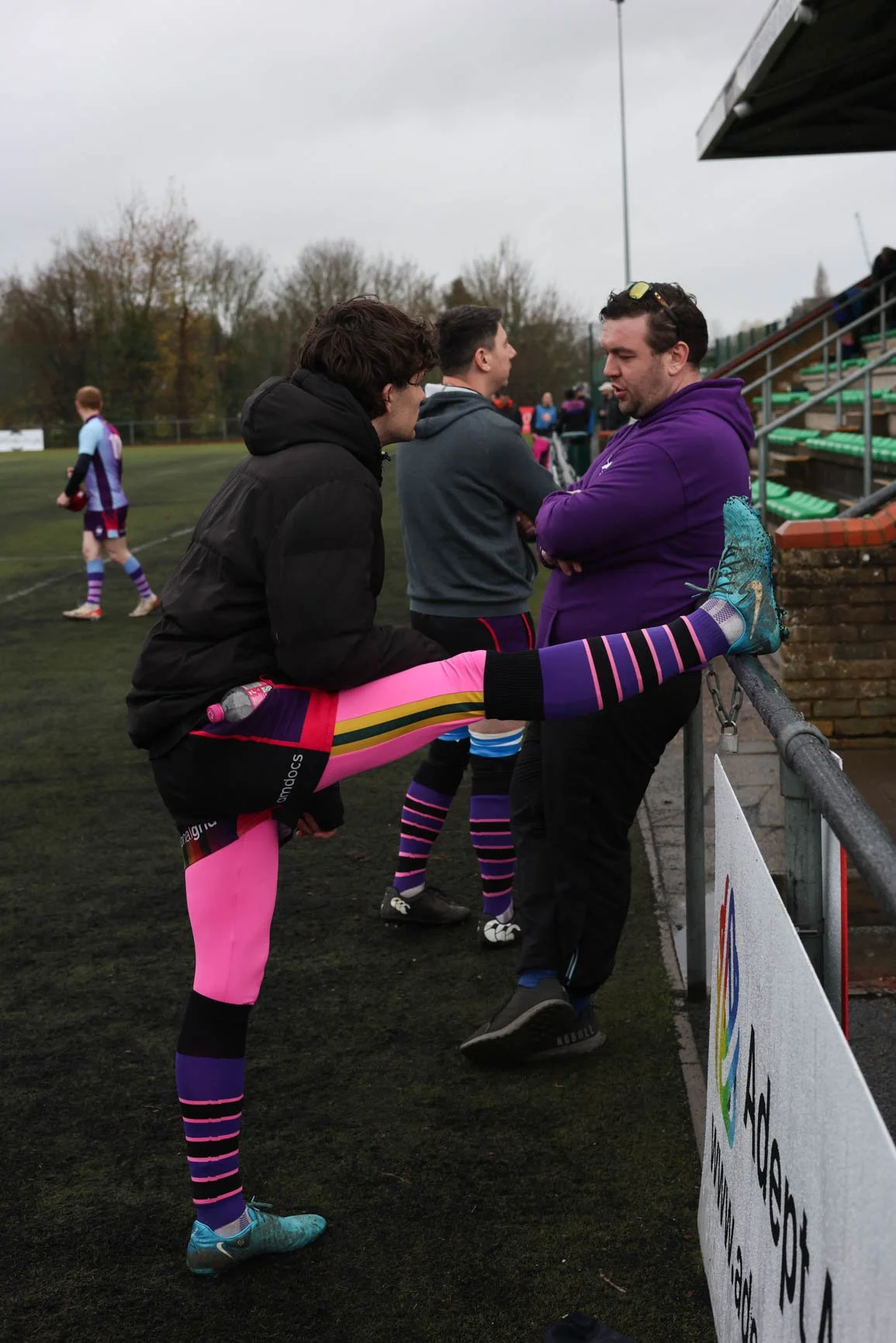 A female rugby player stretching her leg on a fence while talking to a man at a rugby field. The player is wearing pink and purple striped socks, pink shorts, a black jacket, and blue shoes. Other players and people are in the background on a cloudy 