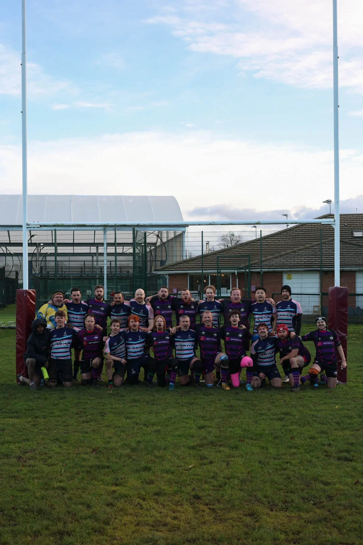 Group of rugby players and supporters on a rugby field posing for a team photo, with goalposts and a sports facility in the background.