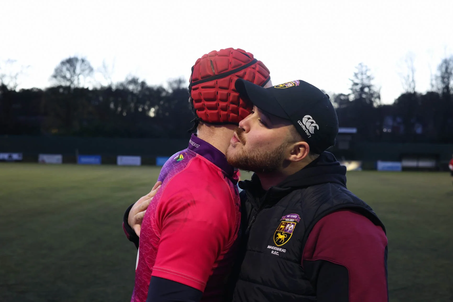 Two men are sharing an emotional embrace on a sports field during dusk, one wearing a red sports helmet and red sports jersey, and the other in a black vest with patches and a cap.