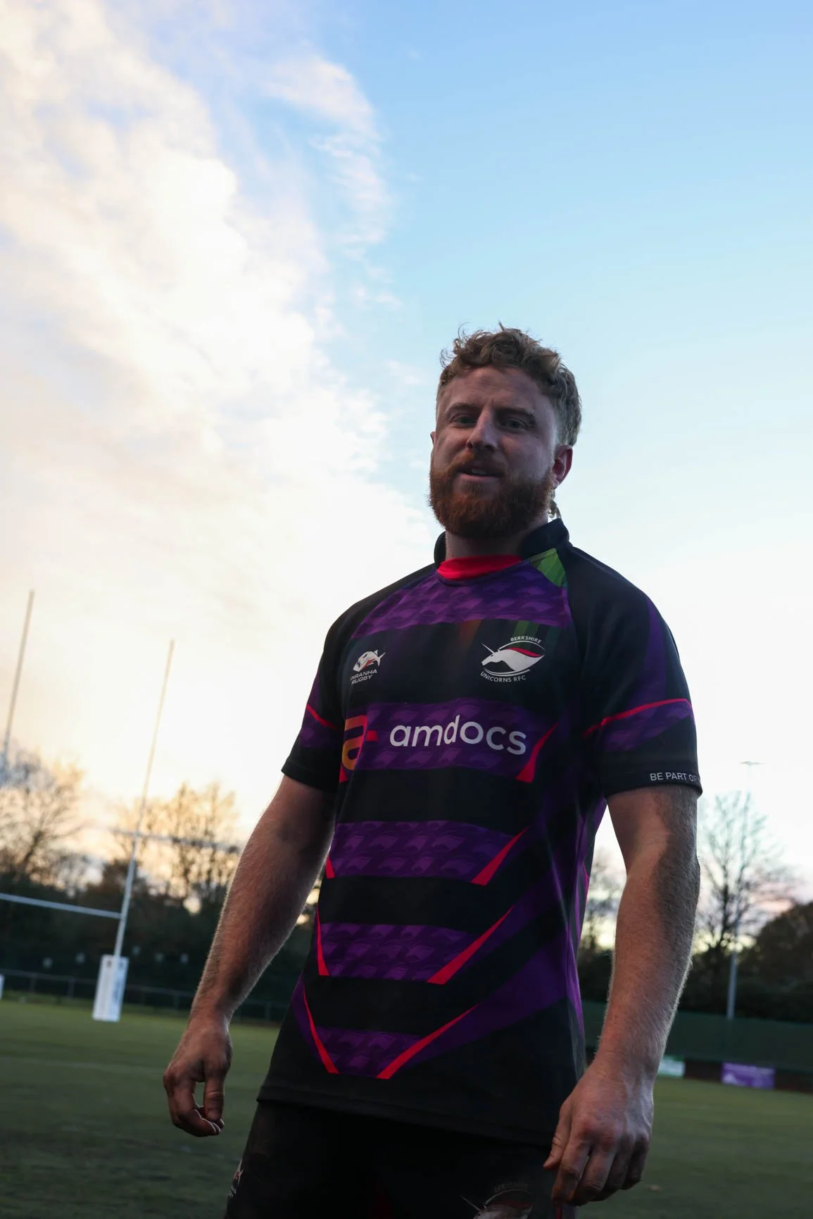 A man with a beard and curly hair wearing a purple and black rugby jersey with sponsor logos, standing on a rugby field during sunset.
