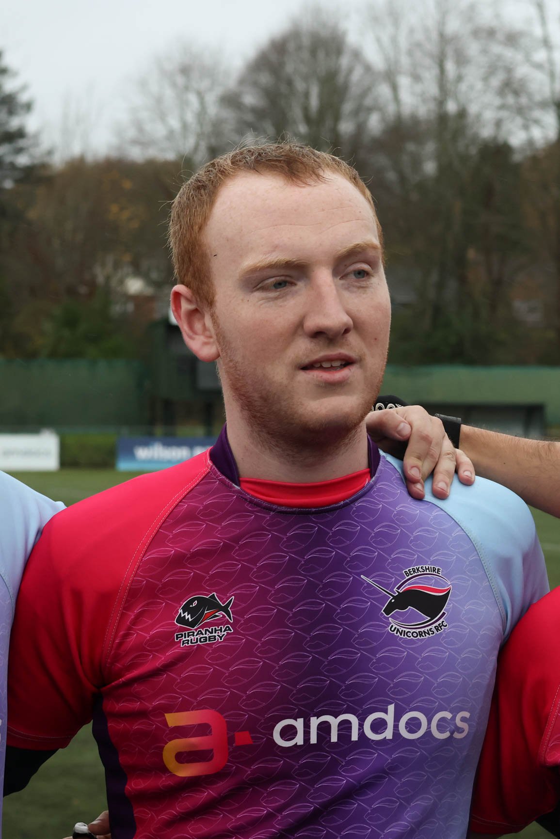 A young male rugby player with red hair and freckles wearing a maroon and purple rugby jersey with logos, standing on a rugby field with trees in the background.