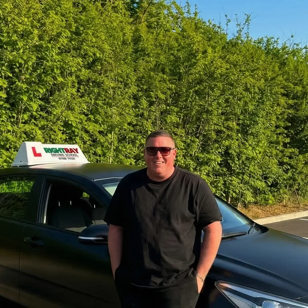 Man standing in front of a black driving school car outdoors on a sunny day, wearing sunglasses and a black T-shirt, with green bushes and a blue sky in the background.