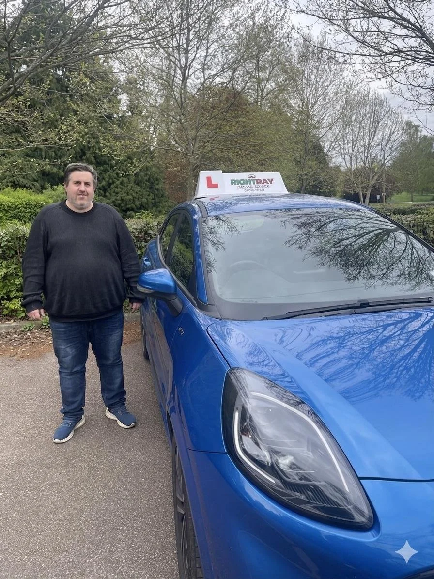 Man standing next to a blue driving school car with a 'L' sign on top, in an outdoor parking area surrounded by trees.