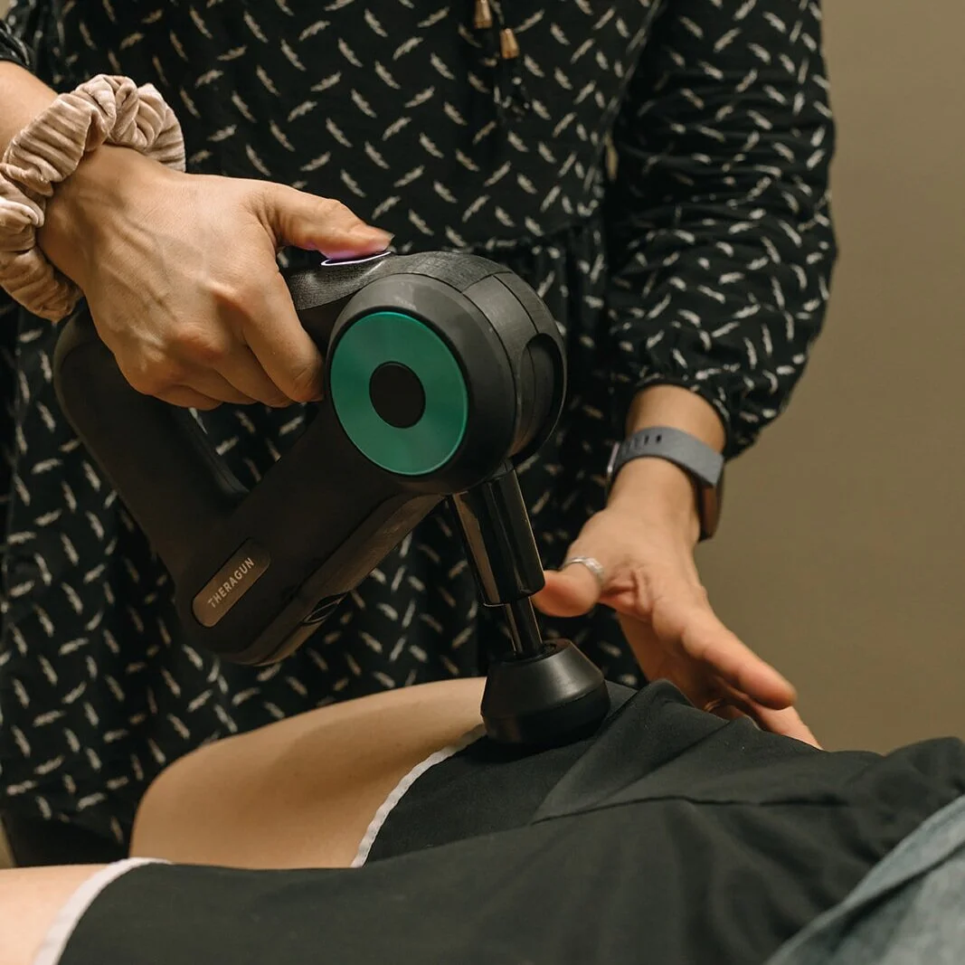 A person receiving physical therapy treatment with a Theragun device on their thigh, while a therapist applies pressure with their hand.