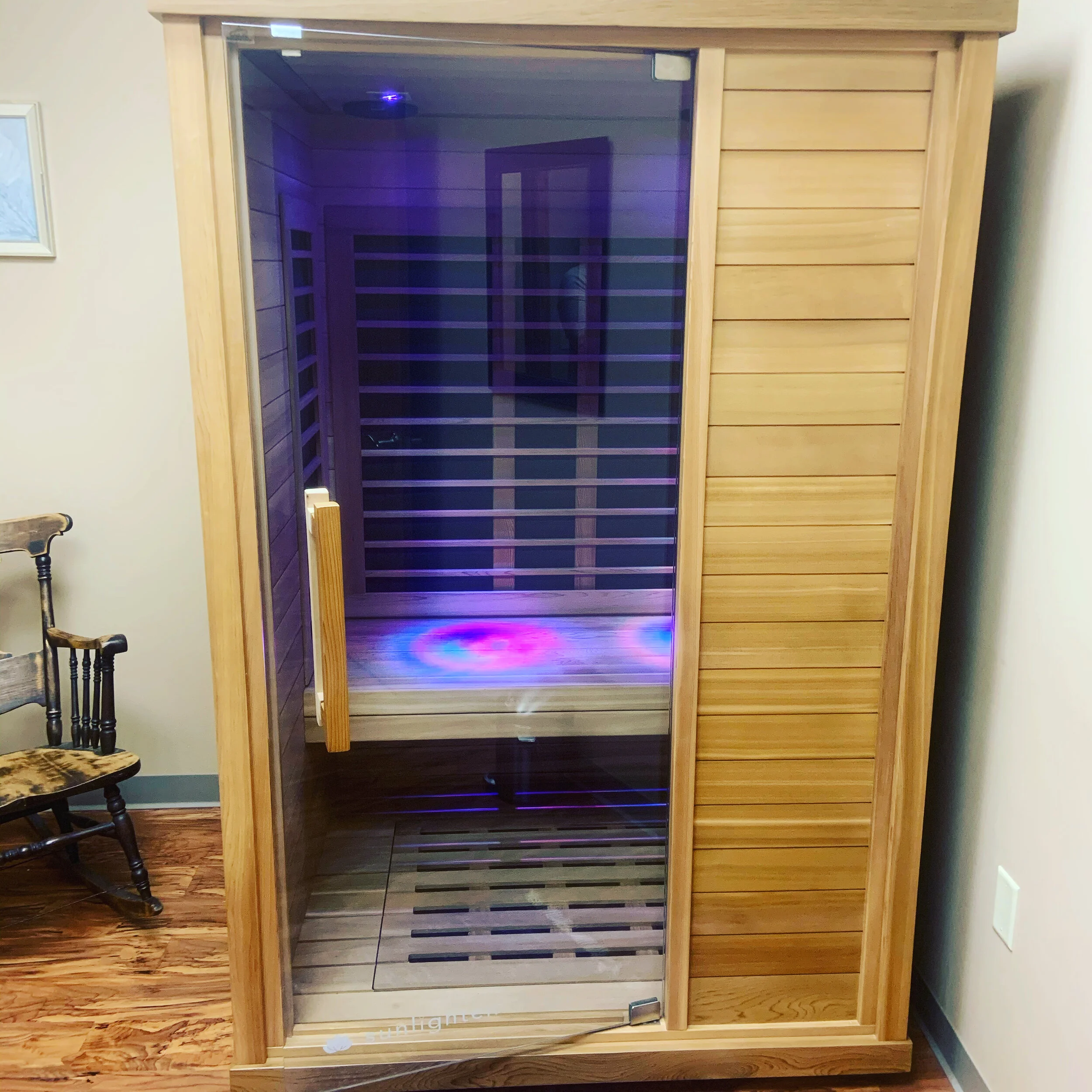 A wooden cabinet with glass doors, housing a wine rack inside, illuminated by purple LED lights, located in a room with hardwood flooring and a chair nearby.