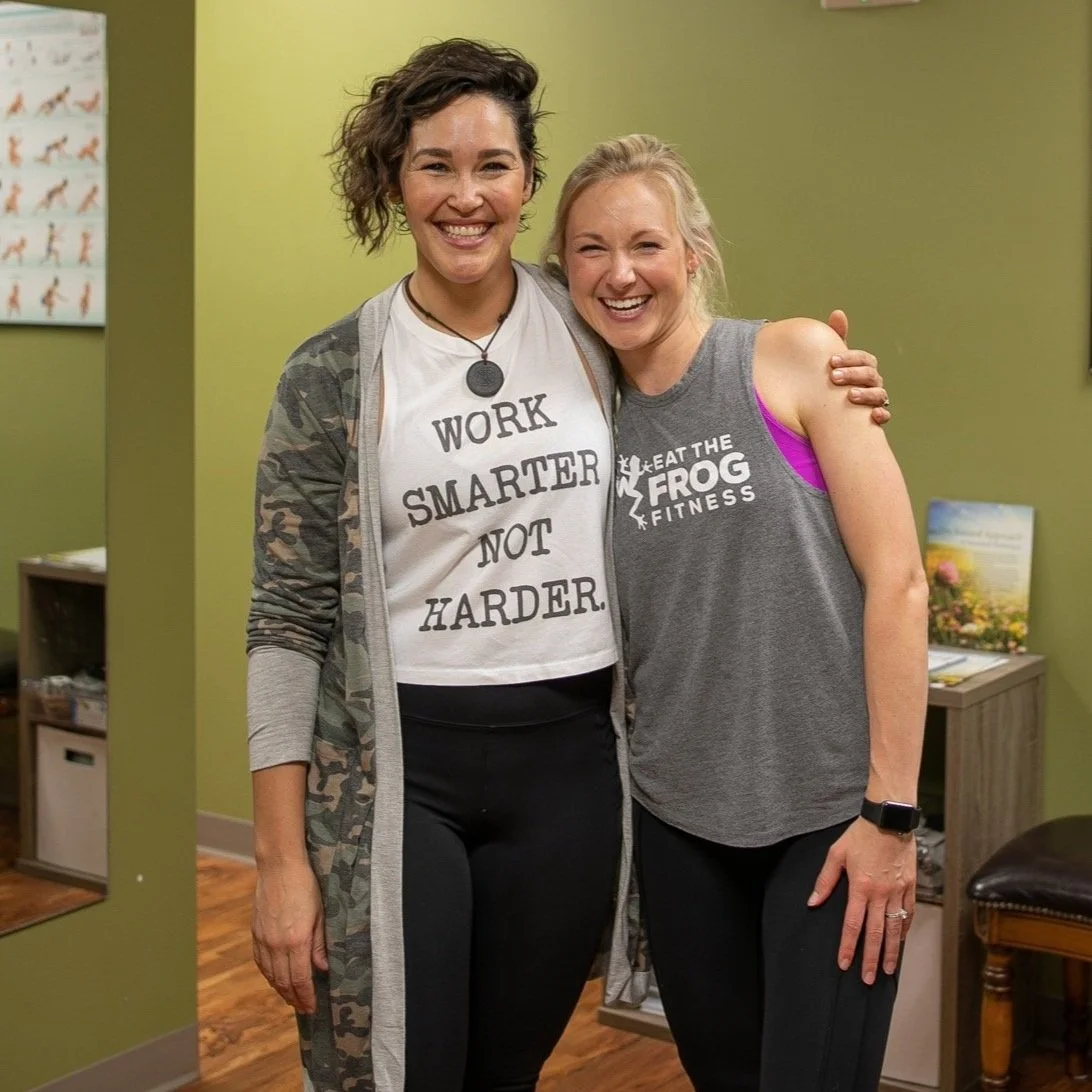 Two women smiling and hugging in a fitness center. One is wearing a white t-shirt with the text 'WORK SMARTER NOT HARDER' and a camouflage jacket. The other wears a gray tank top with 'EAT THE FROG FITNESS' written on it.