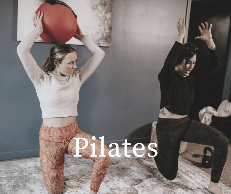 Two women practicing Pilates indoors, one holding a large red exercise ball above her head, the other mid-jump with arms raised.