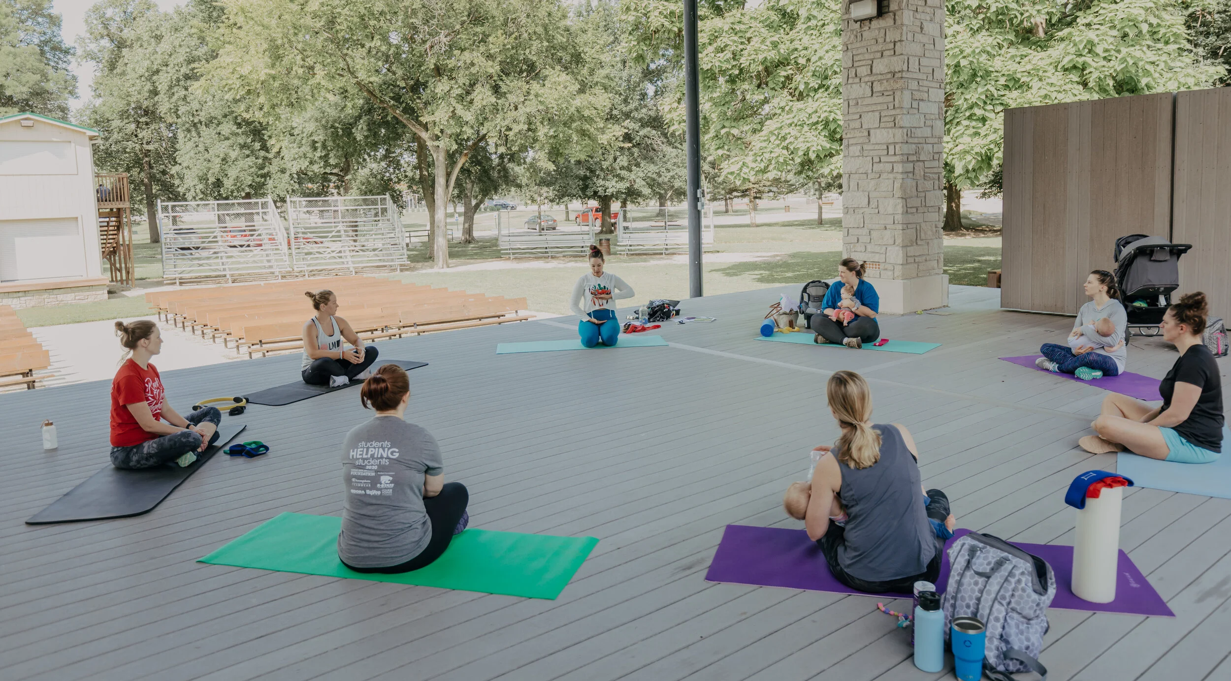 Group of women and children participating in an outdoor yoga or fitness class under a pavilion, sitting on yoga mats with trees and a park in the background.