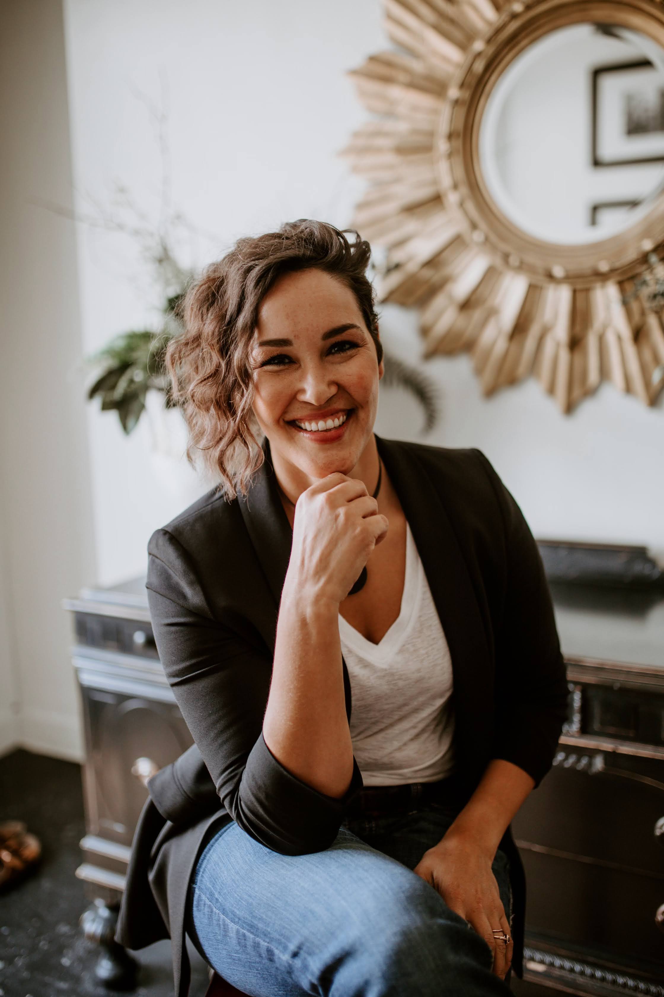 A woman with short, wavy brown hair smiling and sitting in a room with a large round mirror on the wall behind her, wearing a black blazer over a white t-shirt and blue jeans.