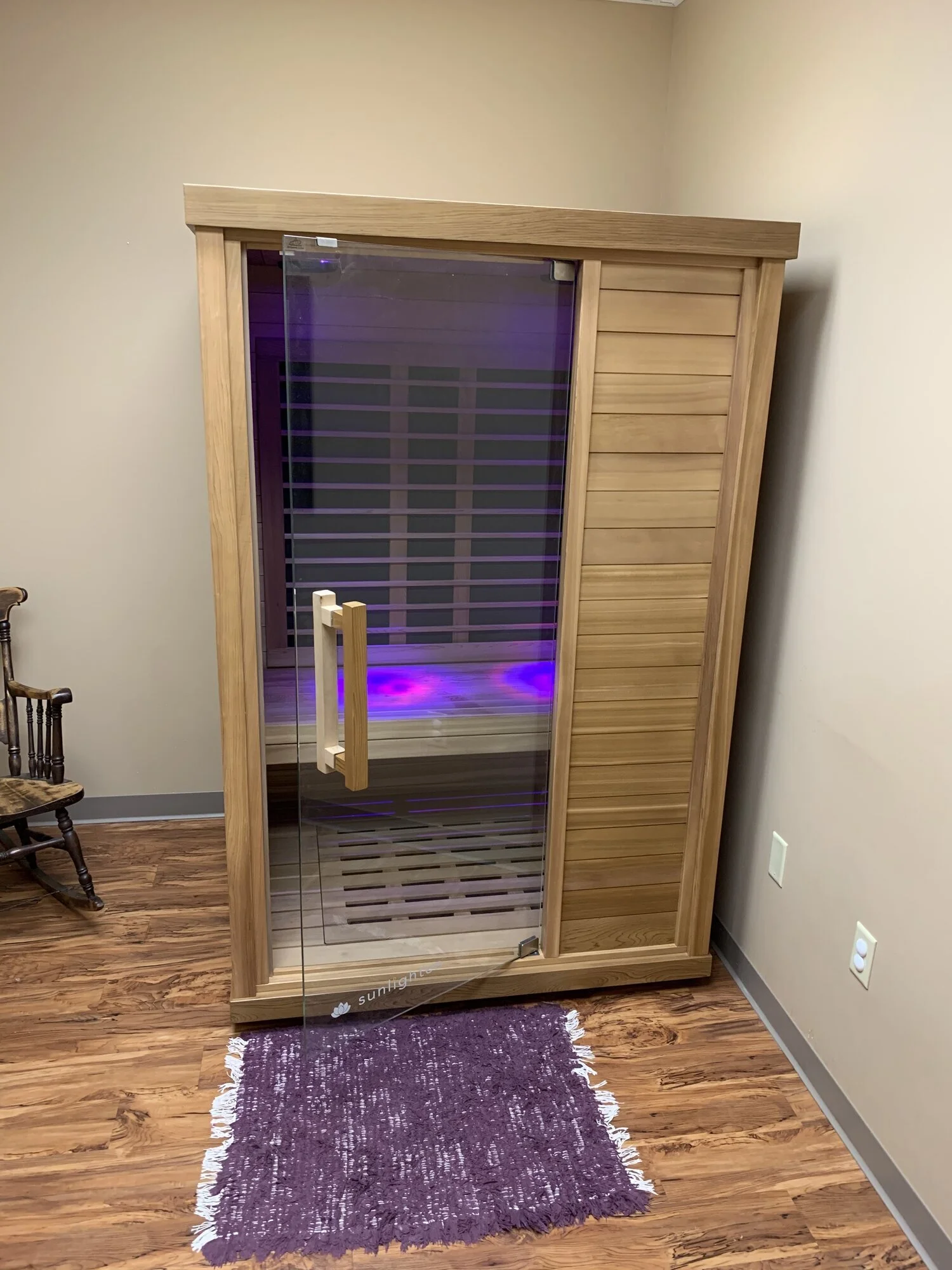 A wooden infrared sauna with a glass door, placed on a hardwood floor next to a purple woven rug. There is a wooden chair to the left and electrical outlets on the wall to the right.