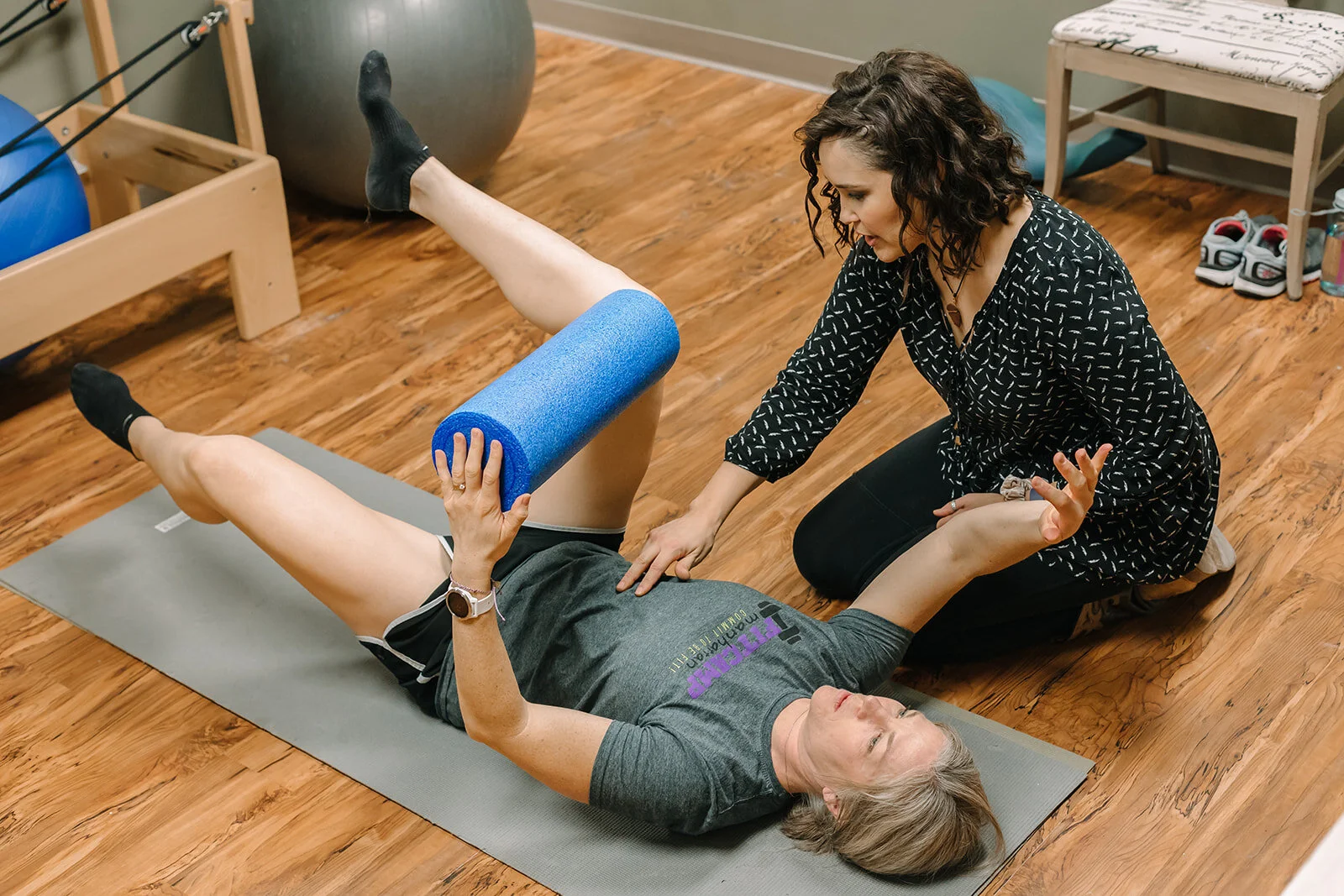 A woman is lying on a gray exercise mat holding a blue foam roller while a trainer kneels beside her, guiding her through a stretch or physical therapy exercise in a gym or therapy room.
