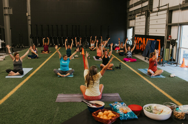 Group of women participating in a yoga class inside a gym, seated on mats with arms raised, instructor leading in front.